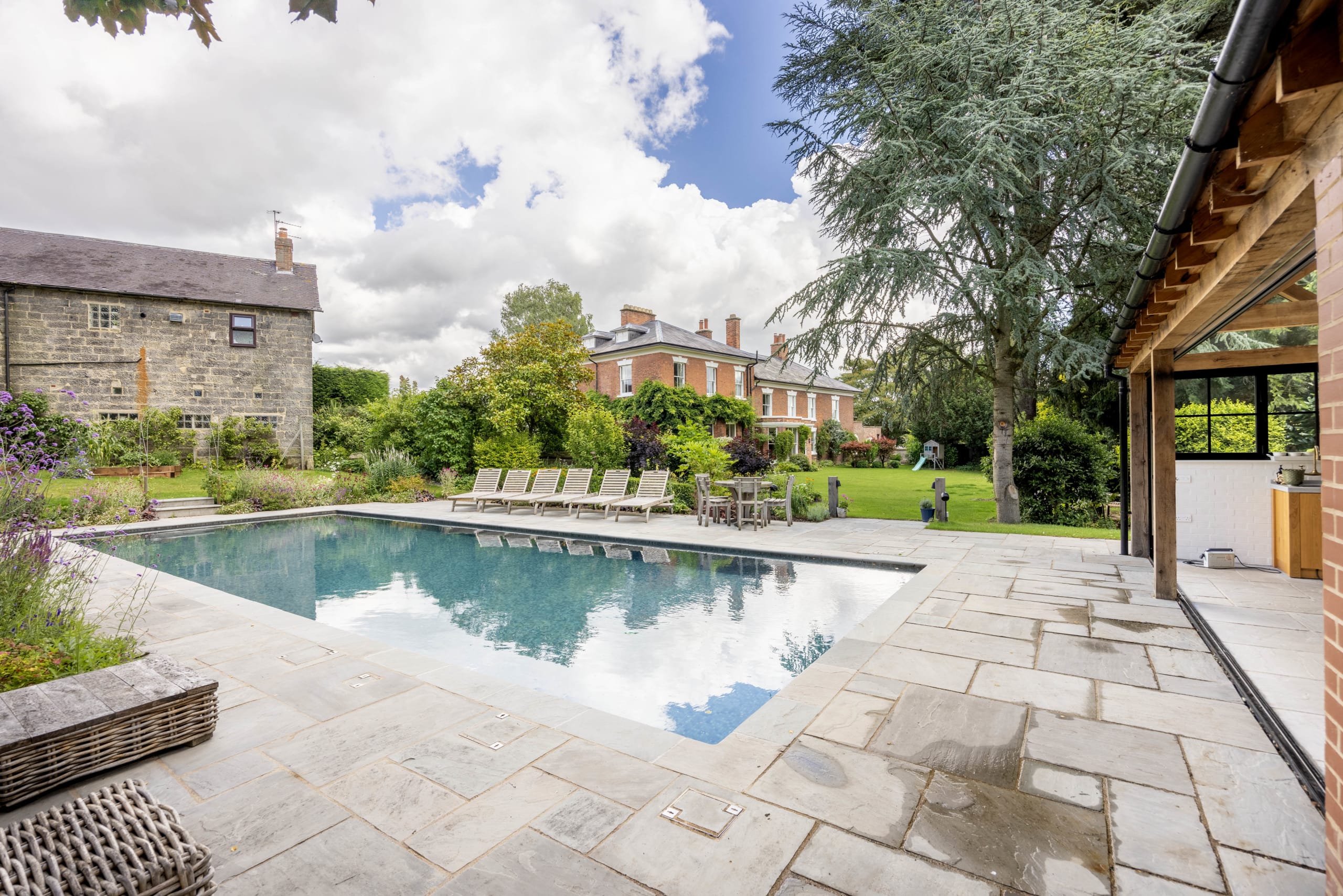 Stone-paved garden with rectangular swimming pool, loungers, and an oak-framed pool house beside a period brick house