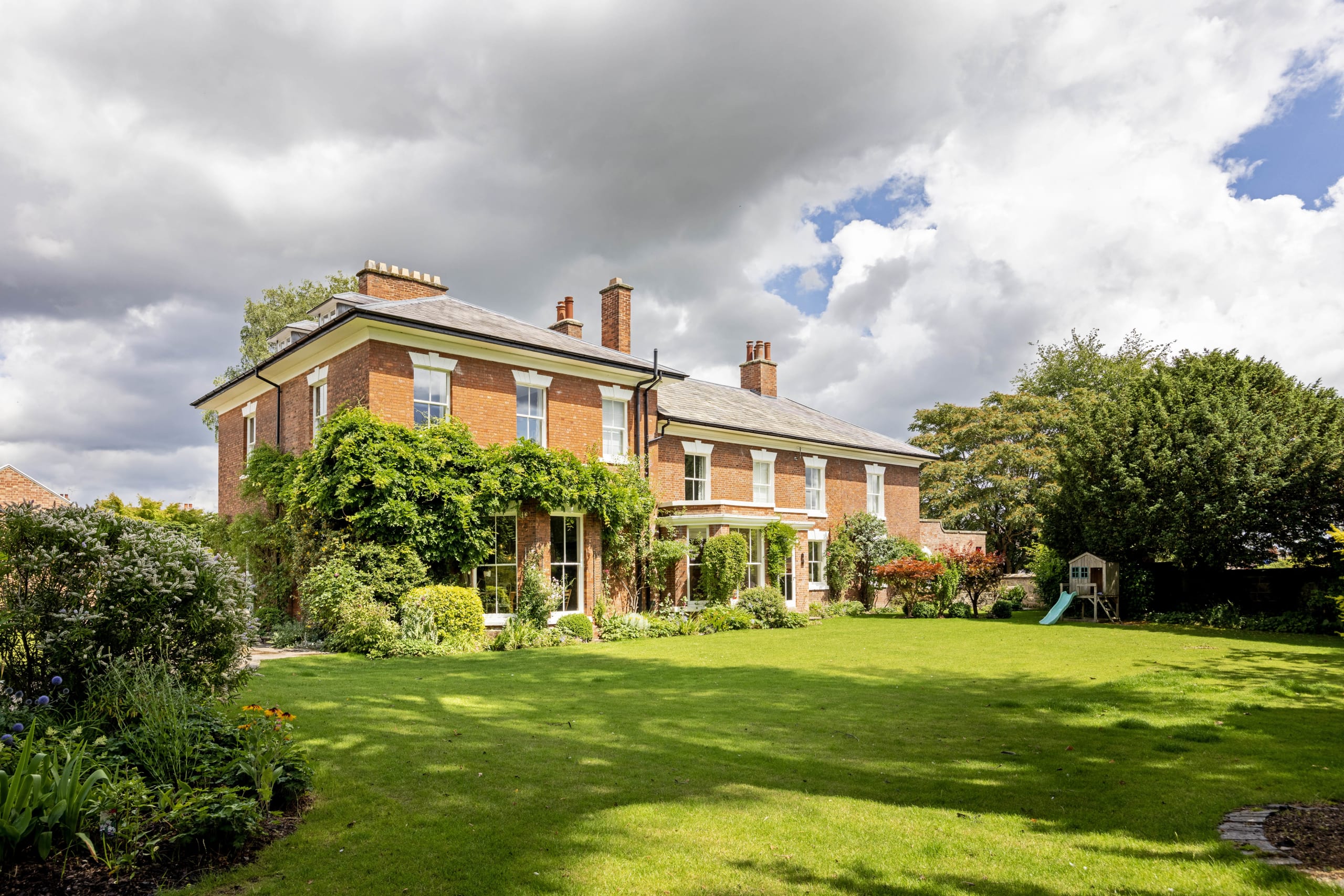 Two-storey brick period house with slate roof, multiple chimneys, and climbing wisteria, seen across a large lawn