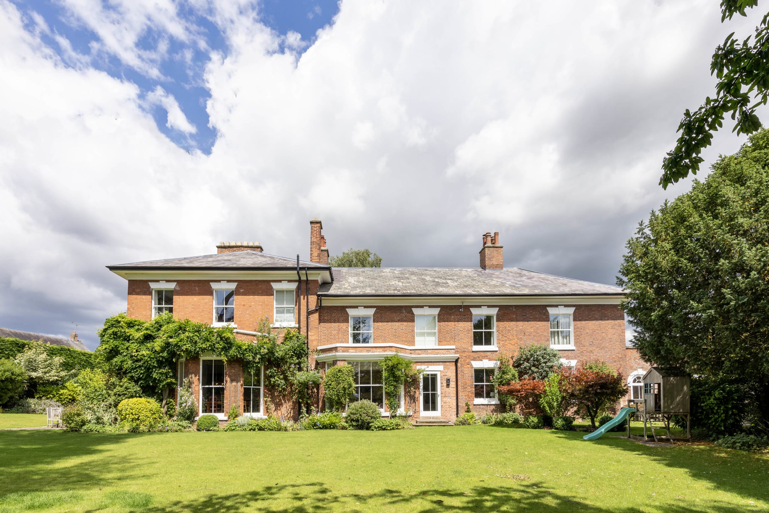 Two-storey brick period house with white sash windows and slate roof, viewed from a landscaped lawn
