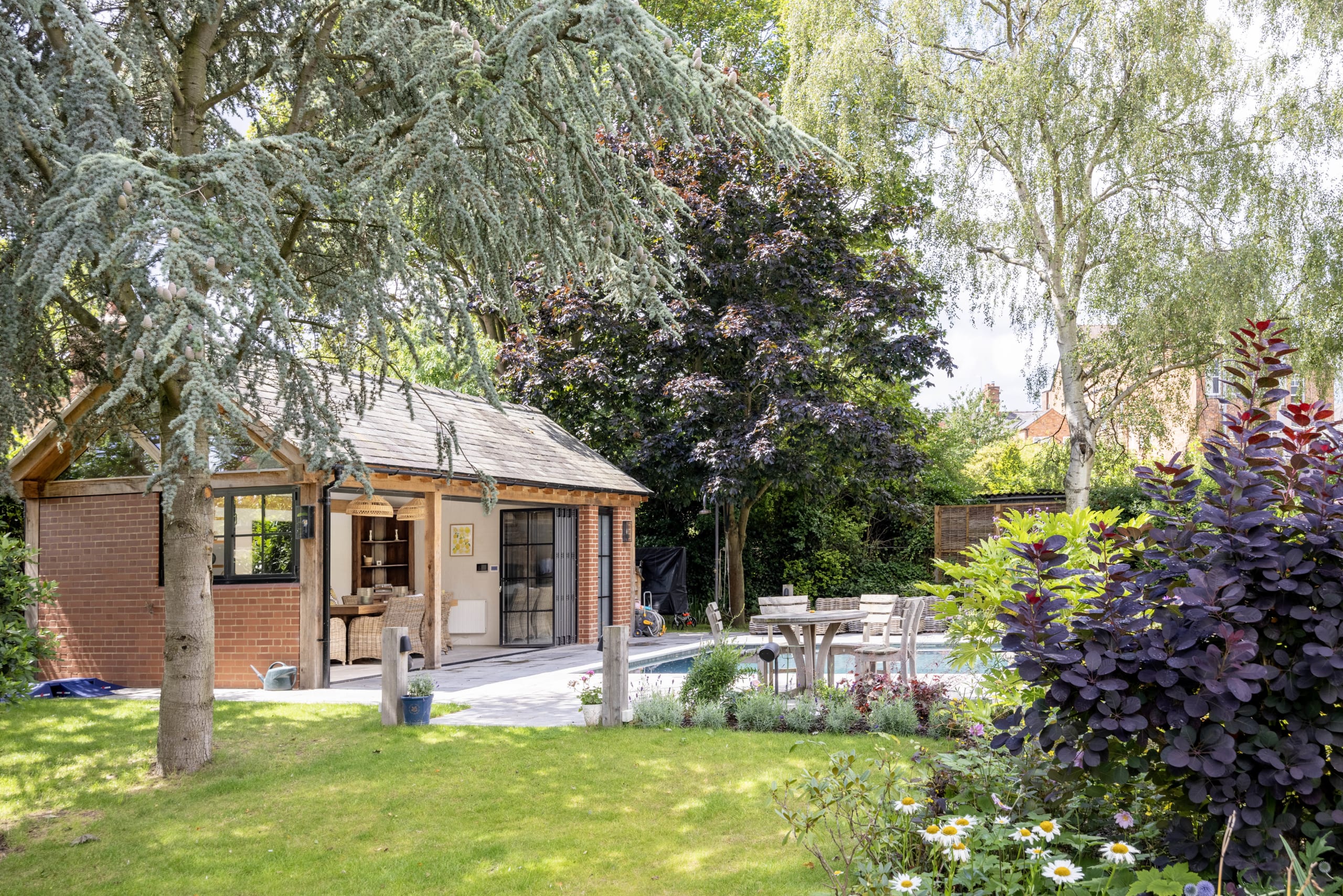 Brick garden pavilion with oak posts and glazed doors opens onto a patio, pool, and lush planted garden