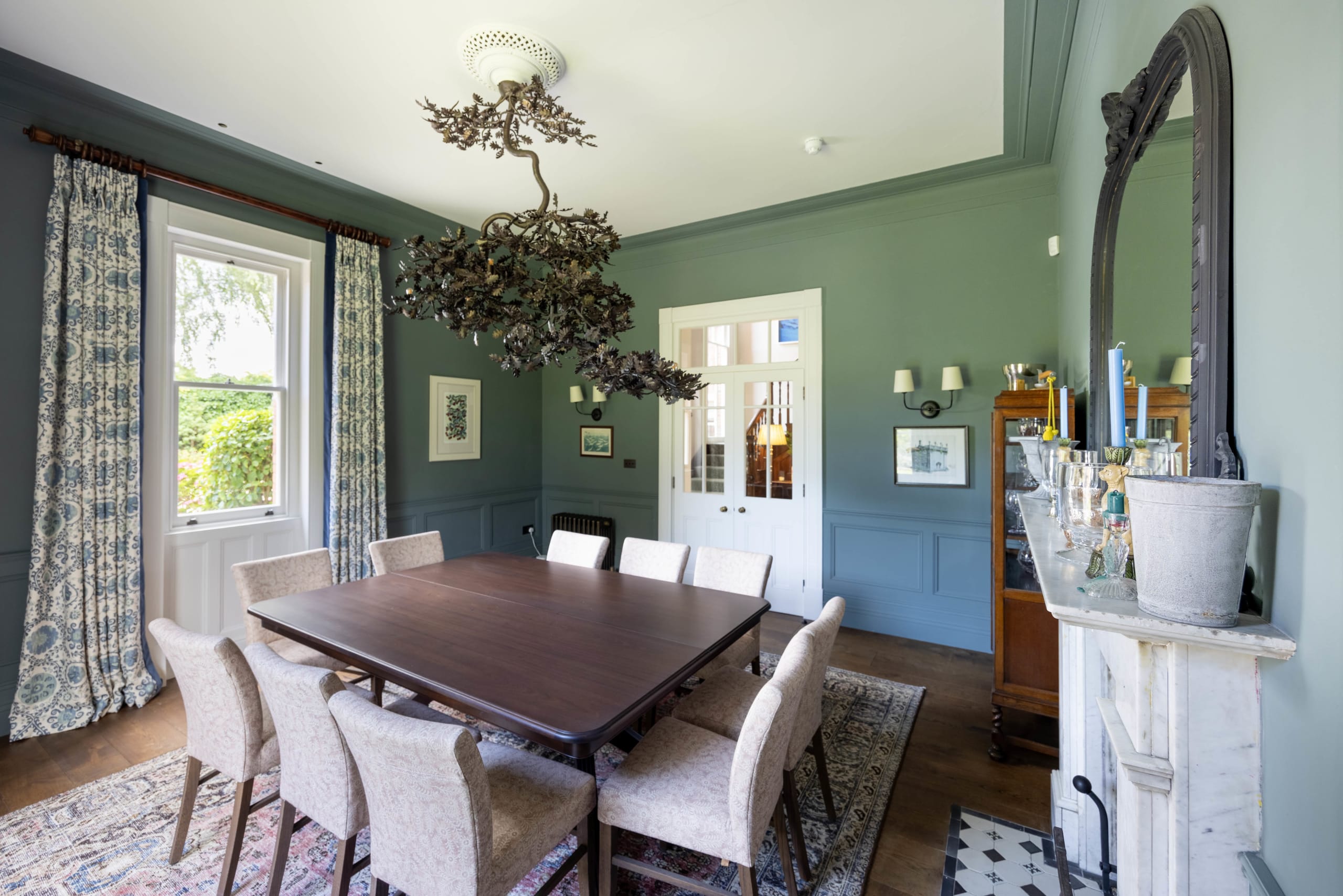Sage-green dining room with dark wood table, upholstered chairs, and an ornate leaf-form chandelier