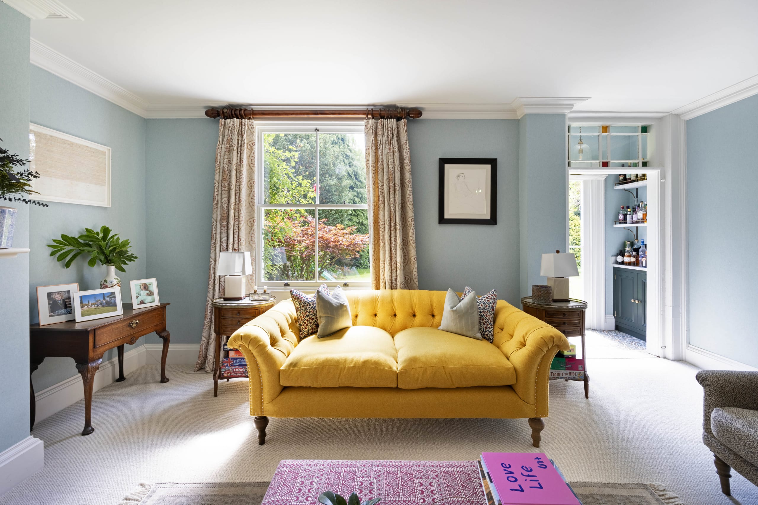 Pale blue traditional living room with a yellow tufted sofa, sash window, and antique side tables