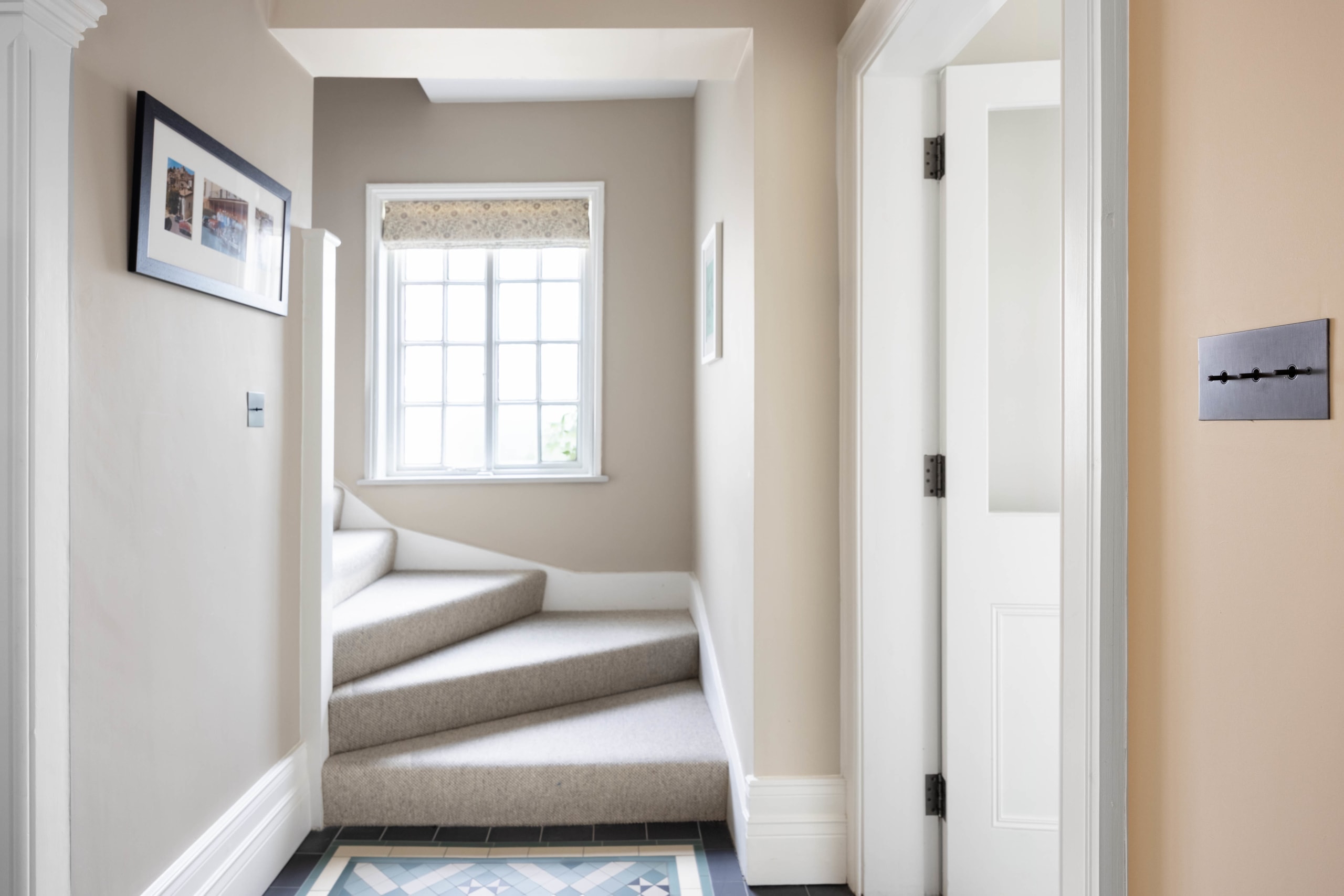 Carpeted staircase turning beside a sash window in a softly painted hallway with patterned floor tiles