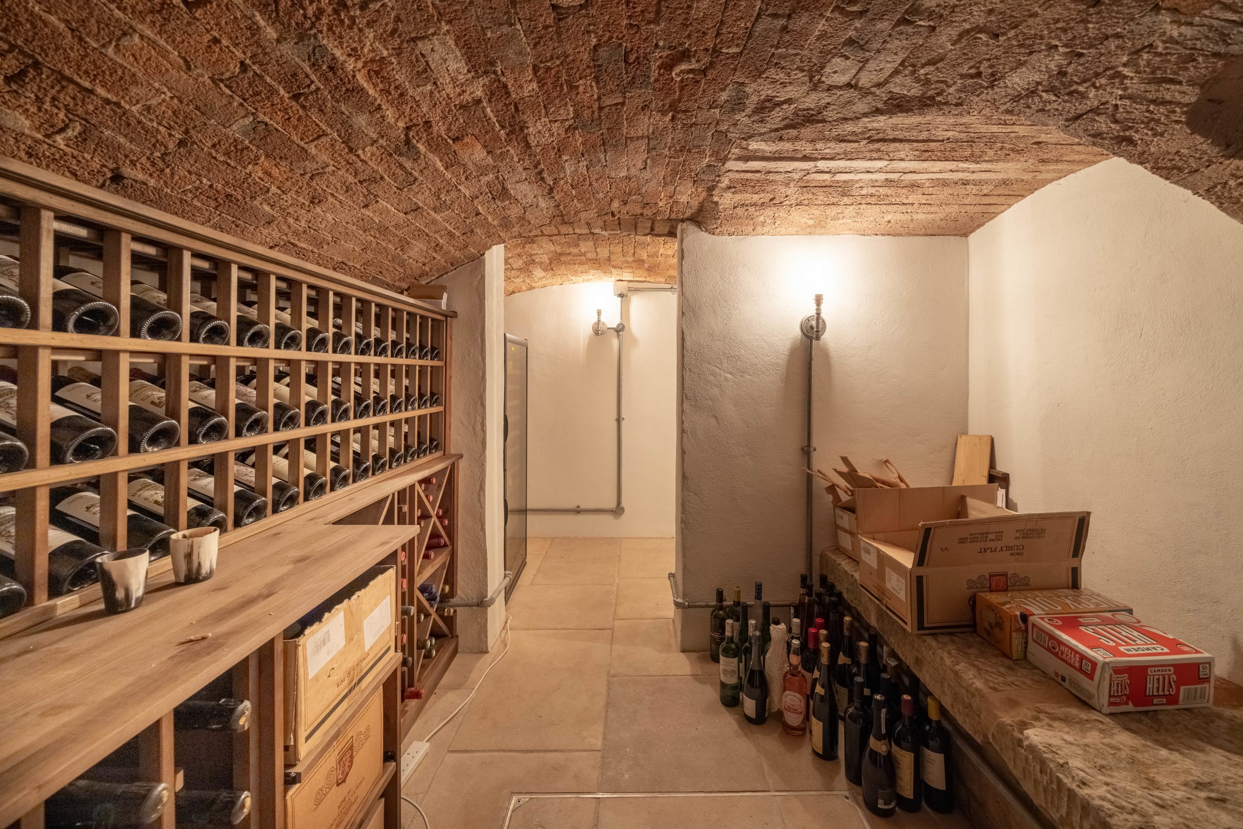 Vaulted brick wine cellar with wooden bottle racks and stone floor lined with bottles and crates