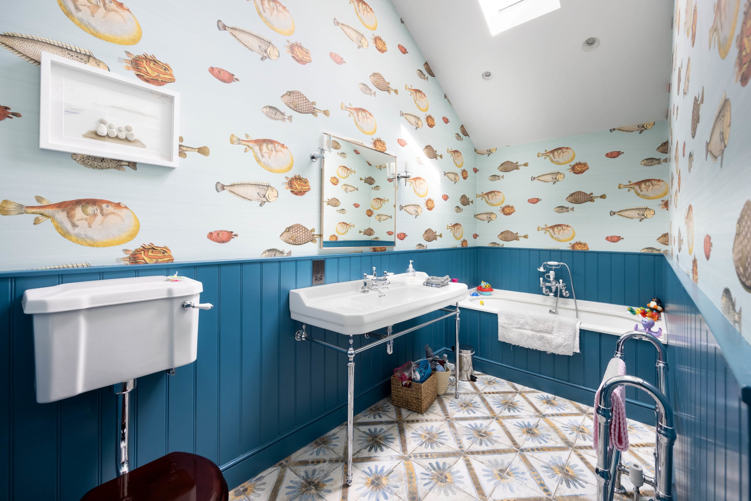 Attic bathroom with fish-print wallpaper, blue wainscoting, and patterned tile floor beneath a sloped skylight ceiling