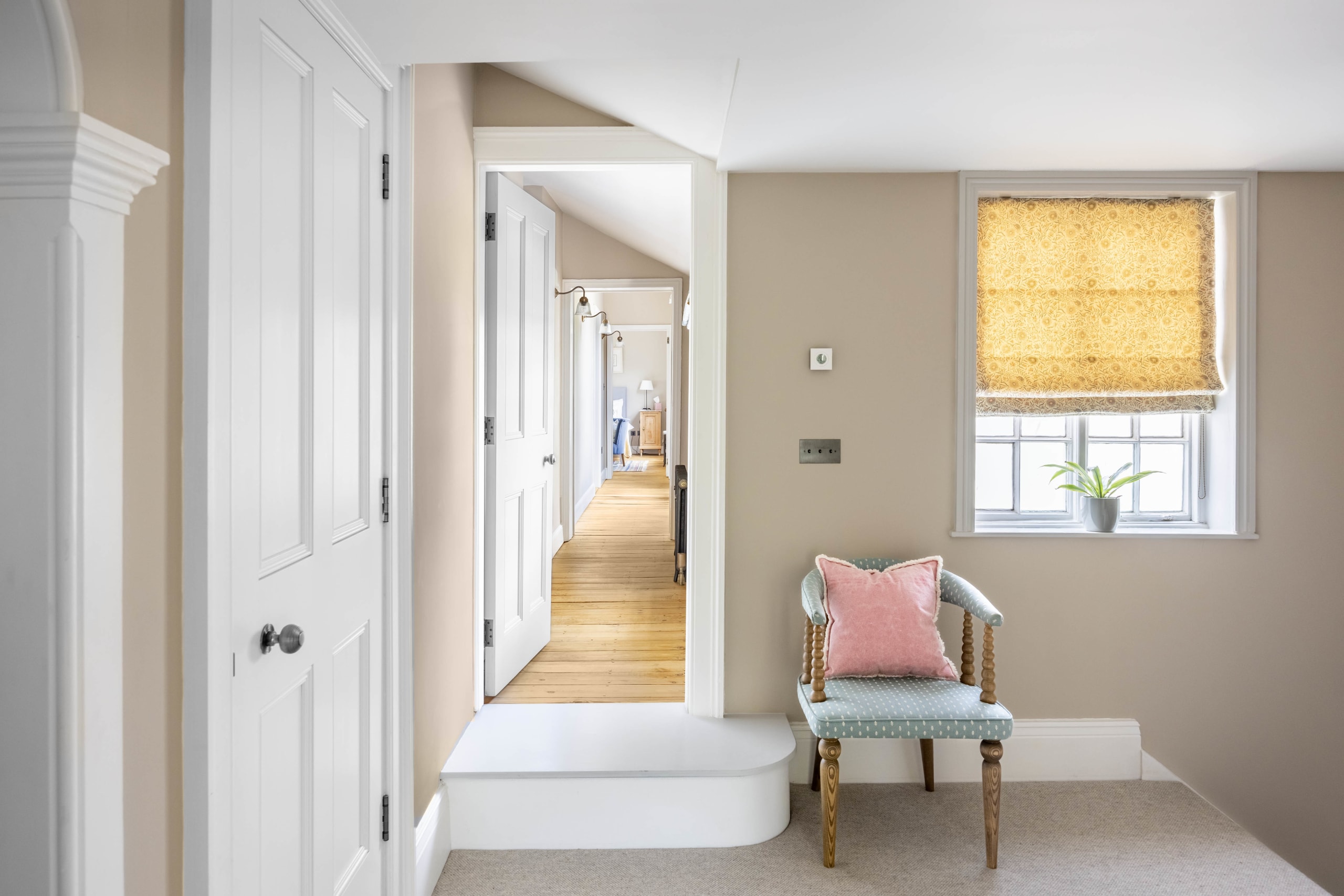 Sunlit hallway with white paneled doors, wood floor, and a striped chair beneath a yellow patterned roman blind