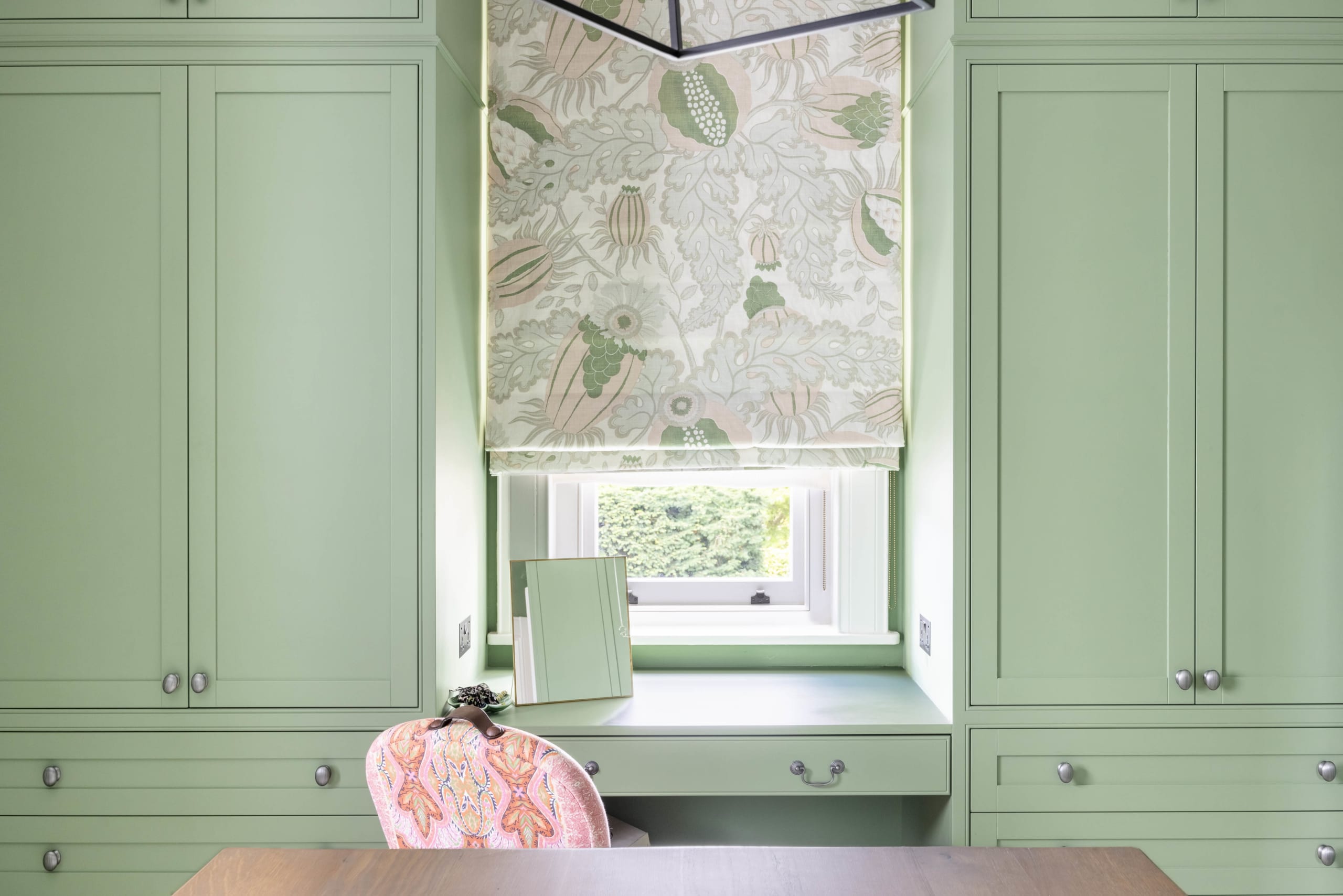 Built-in sage green cabinetry with desk nook featuring botanical Roman shade and pink patterned chair