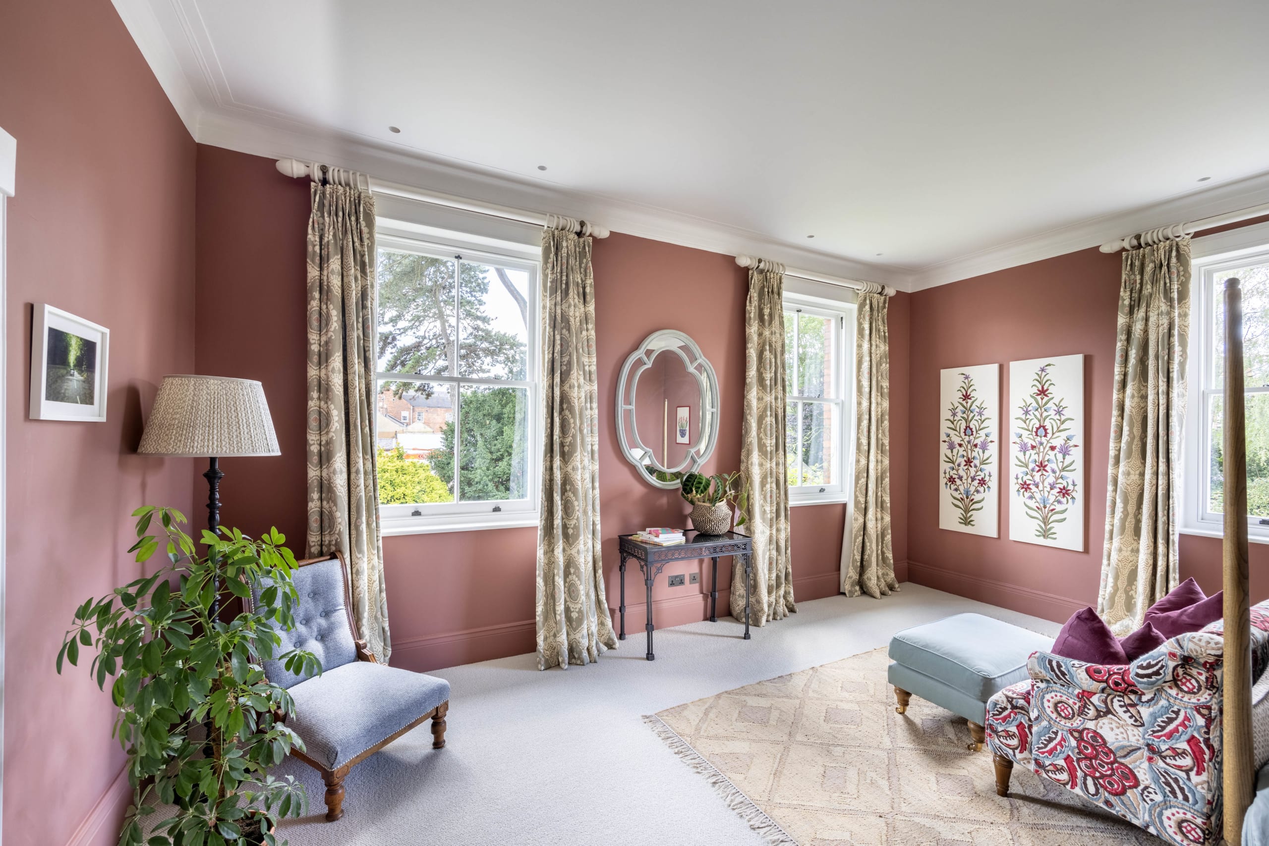 Elegant bedroom with terracotta walls, four-poster bed, blue velvet seating, botanical prints, and tall sash windows