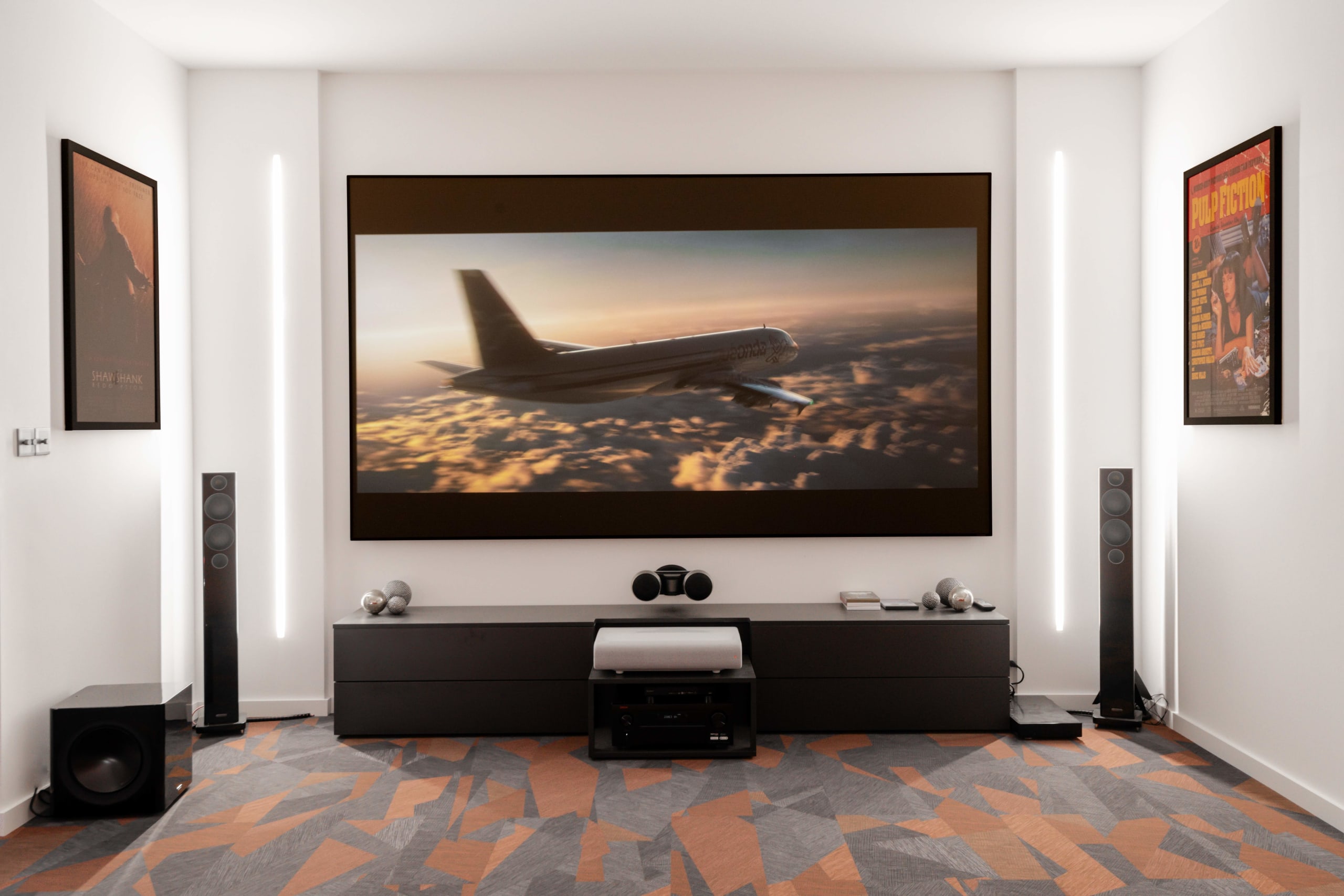 Modern home theater room with large projection screen showing airplane scene, floor speakers, subwoofer, and movie posters