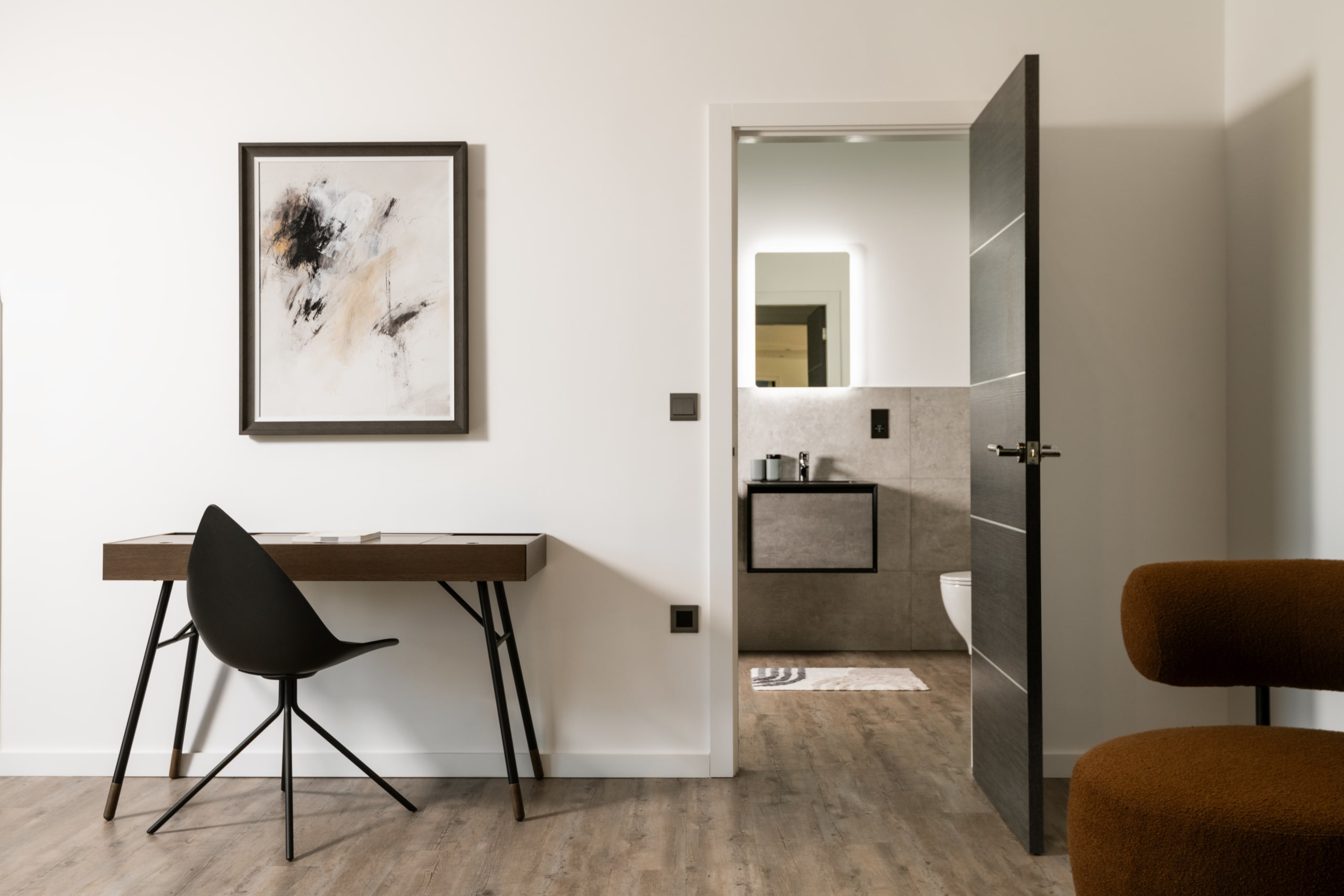 Modern bedroom workspace with minimalist desk, black chair, abstract wall art, and ensuite bathroom with gray tile