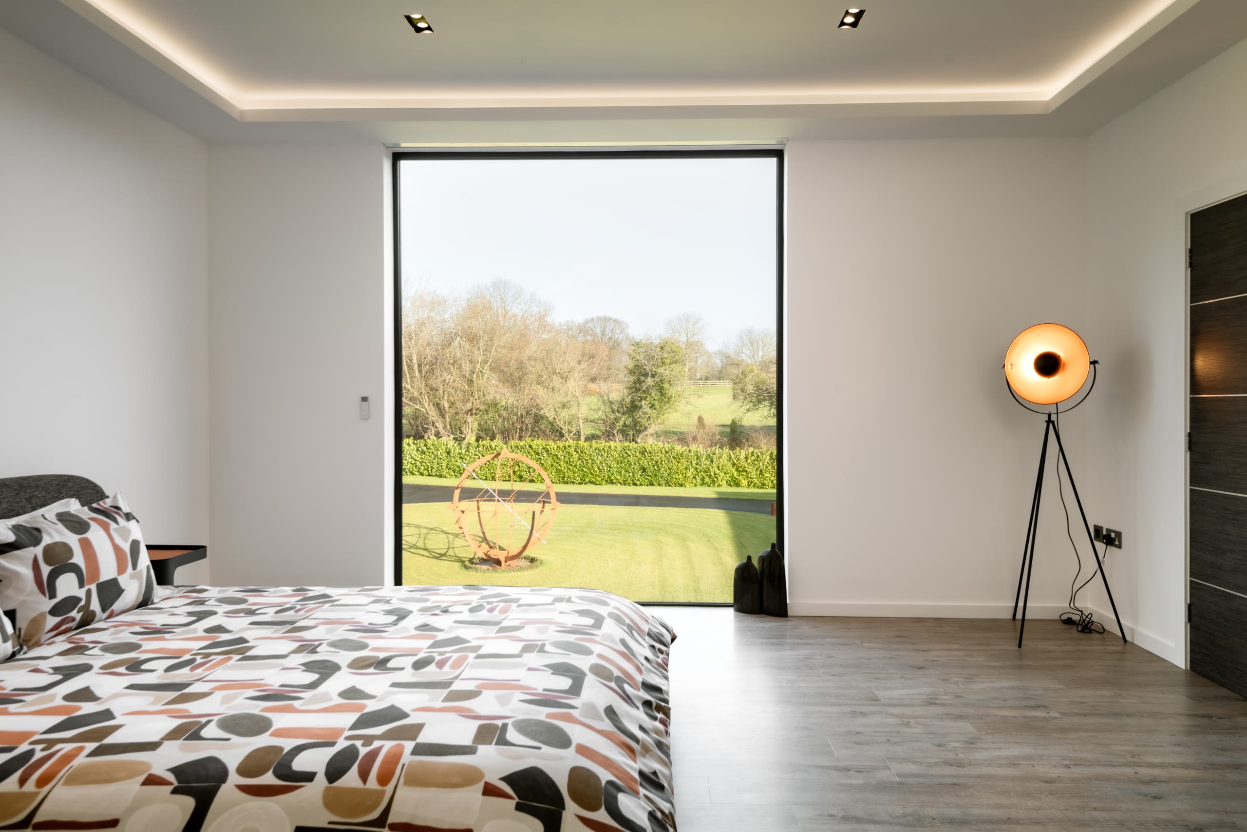 Modern bedroom with geometric bedding, floor-to-ceiling window overlooking garden with metal sculpture, tripod floor lamp