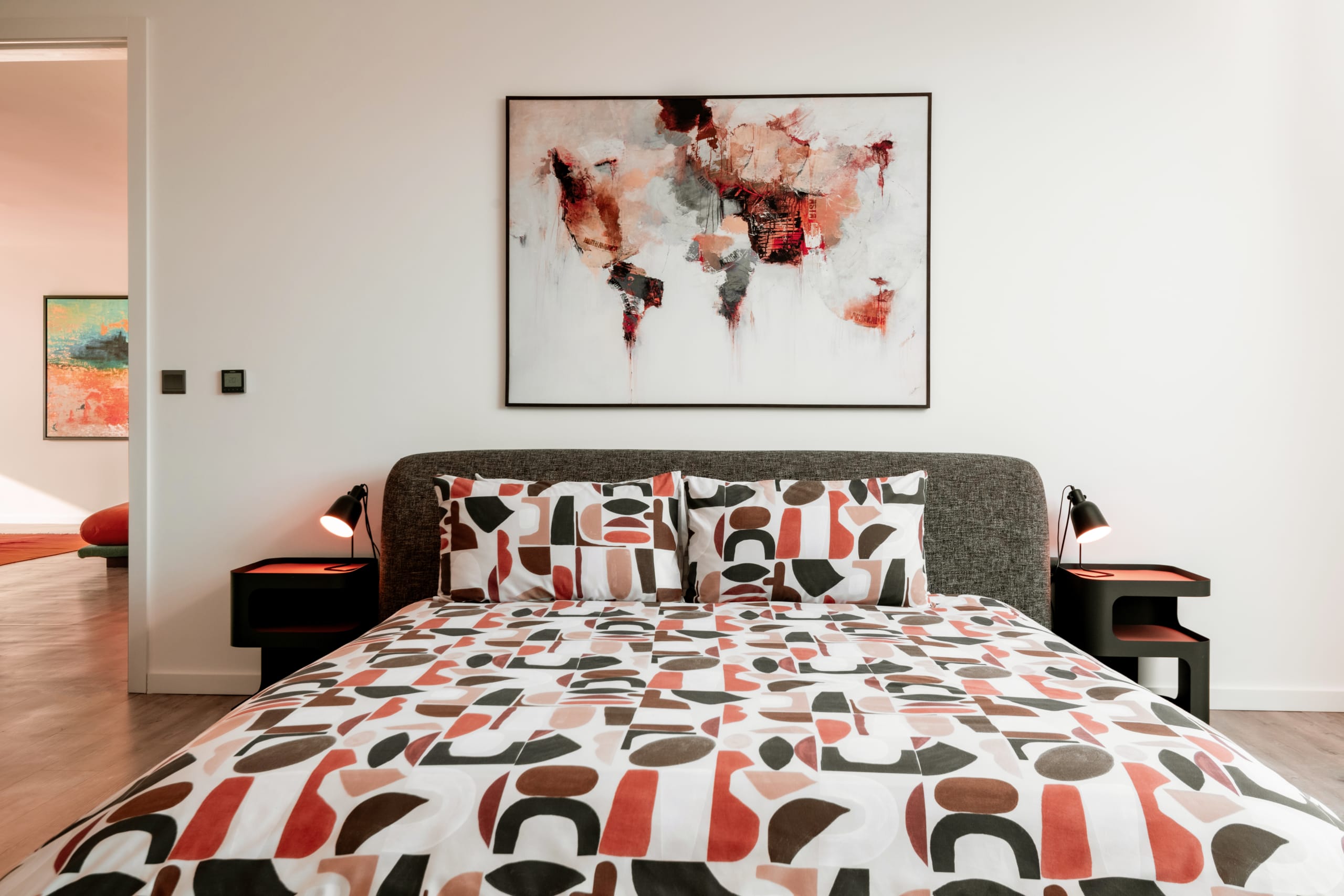 Modern bedroom with geometric patterned bedding, gray upholstered headboard, abstract world map artwork, and black nightstands