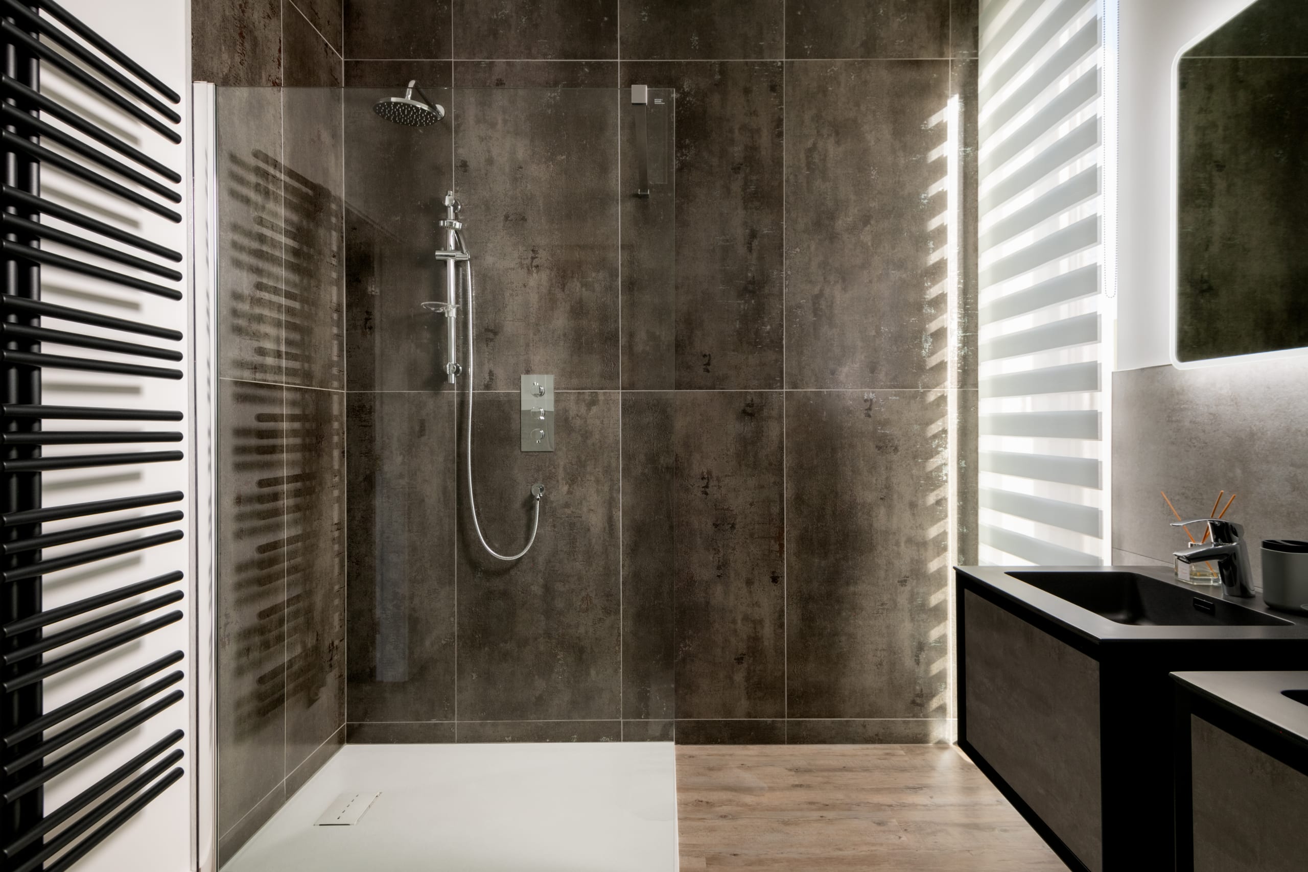 Modern walk-in shower with dark concrete-look tile walls, chrome fixtures, glass panel, and black heated towel rail