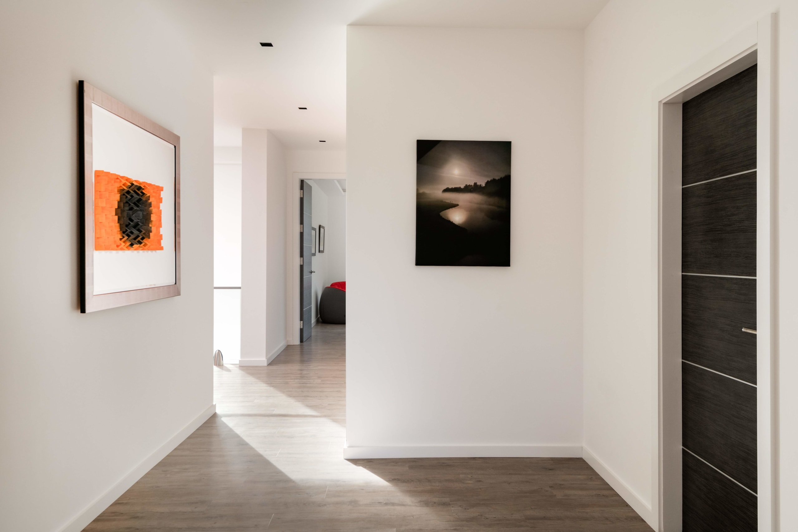 Modern hallway with white walls, wood flooring, geometric orange artwork, black and white landscape photo, and dark wood door