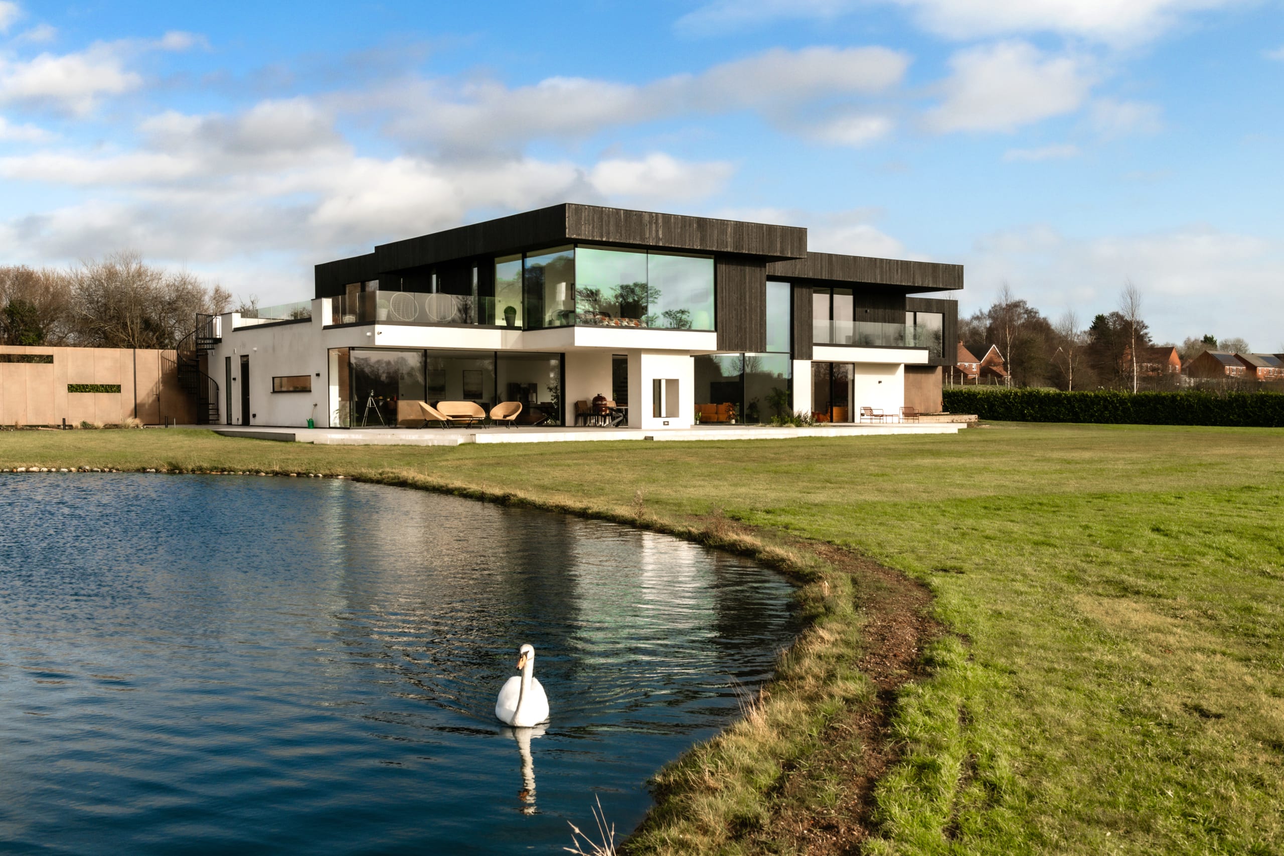 Modern two-storey black and white residence with floor-to-ceiling glass windows overlooking a private pond with swan