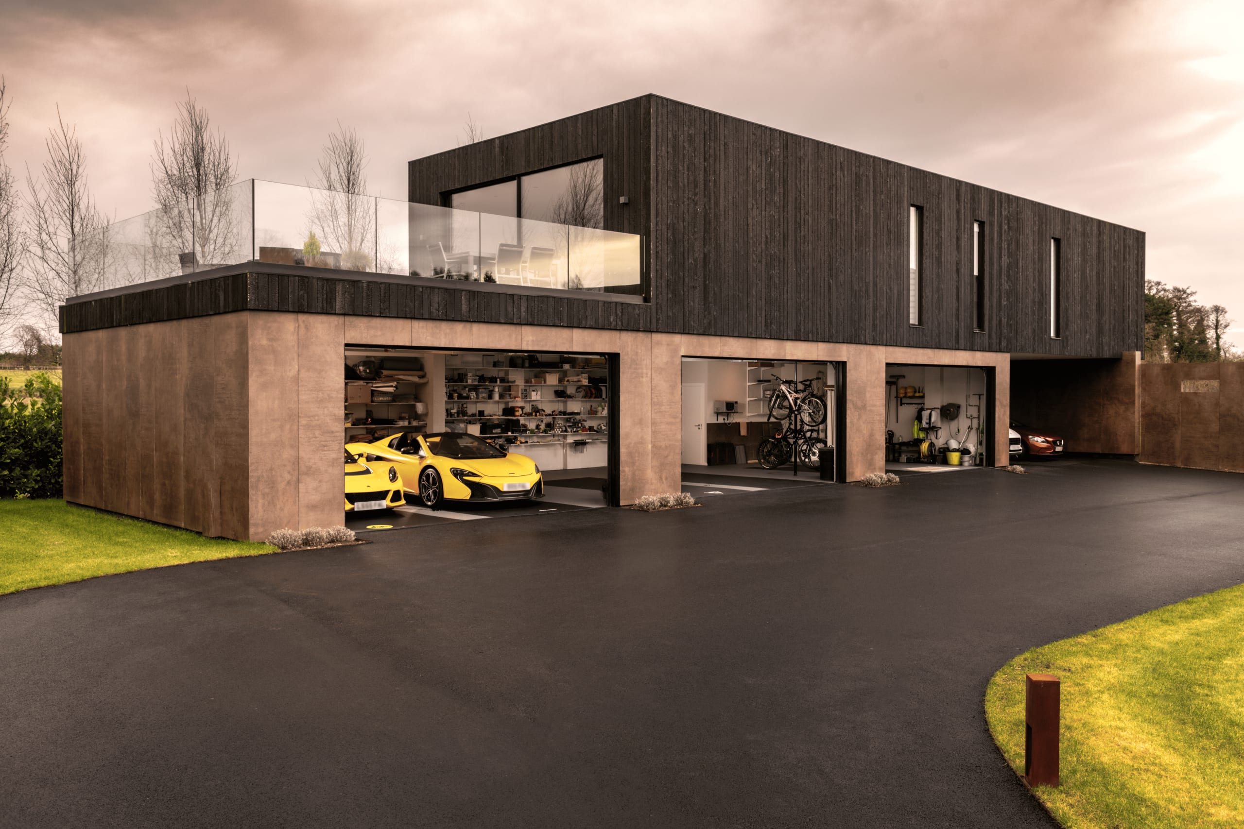 Modern two-story home with dark timber cladding, glass balcony, and four-bay garage featuring yellow McLaren sports car