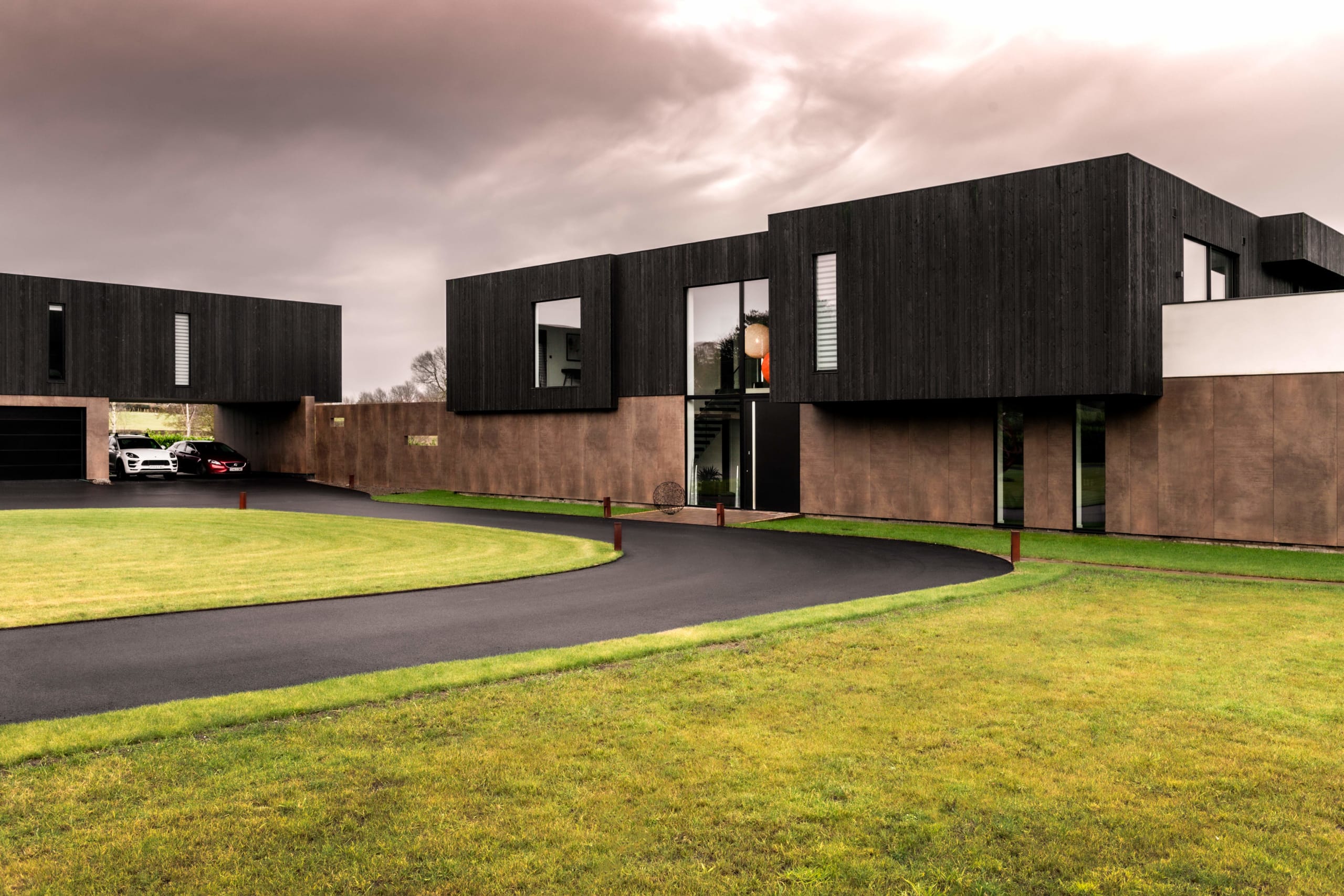Modern two-story home with dark timber cladding, corten steel panels, curved driveway, and manicured lawn with luxury cars