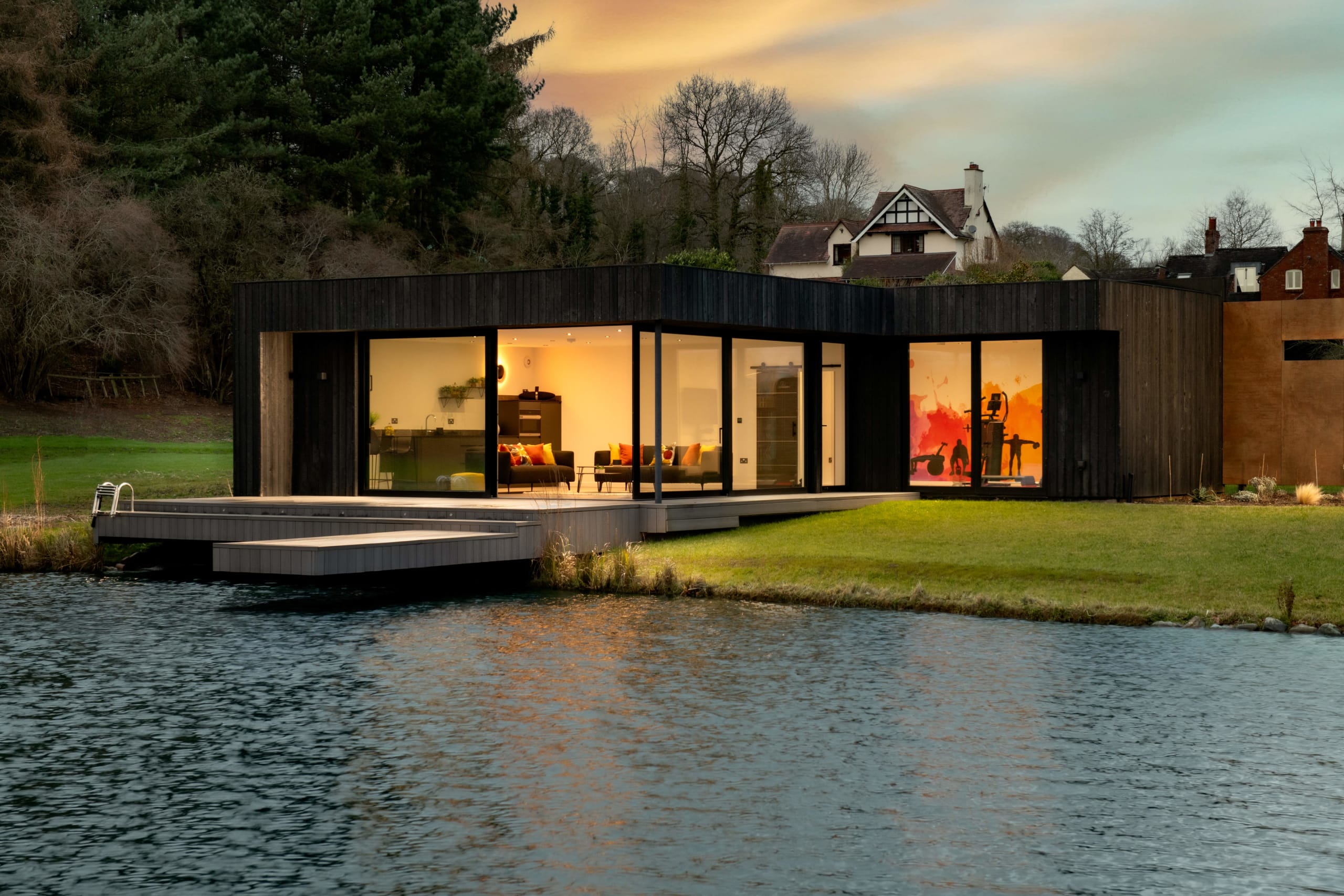 Modern dark timber-clad lakeside pavilion with floor-to-ceiling glass walls, deck over water, illuminated at dusk