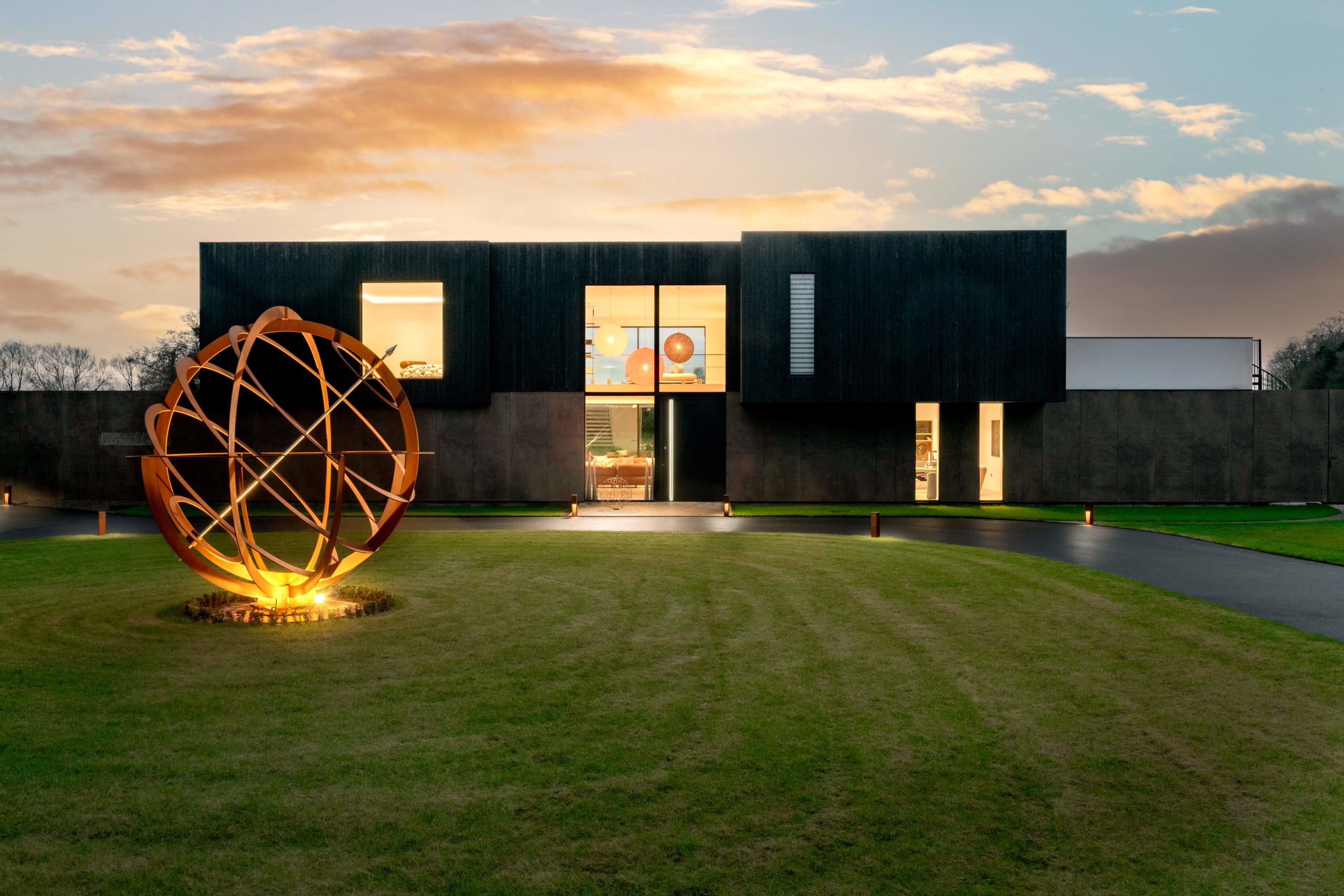 Modern black-clad residence at dusk with illuminated interior, featuring large spherical copper sculpture on manicured lawn