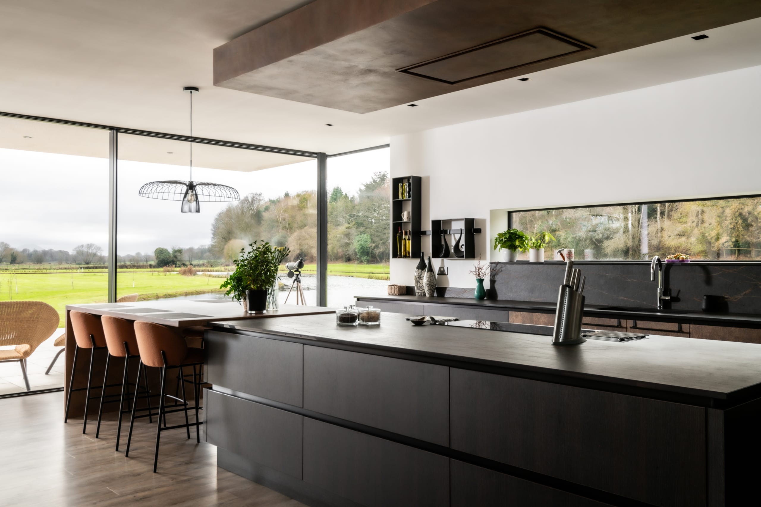 Modern open-plan kitchen with dark cabinetry, breakfast bar seating, and floor-to-ceiling windows overlooking green countryside