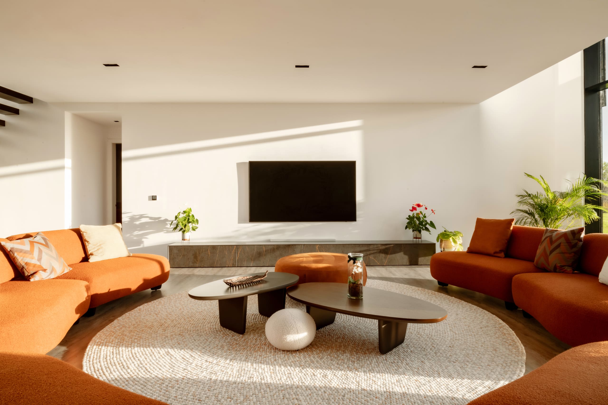 Modern minimalist living room with burnt orange curved sofas, oval coffee tables, round textured rug, and wall-mounted TV