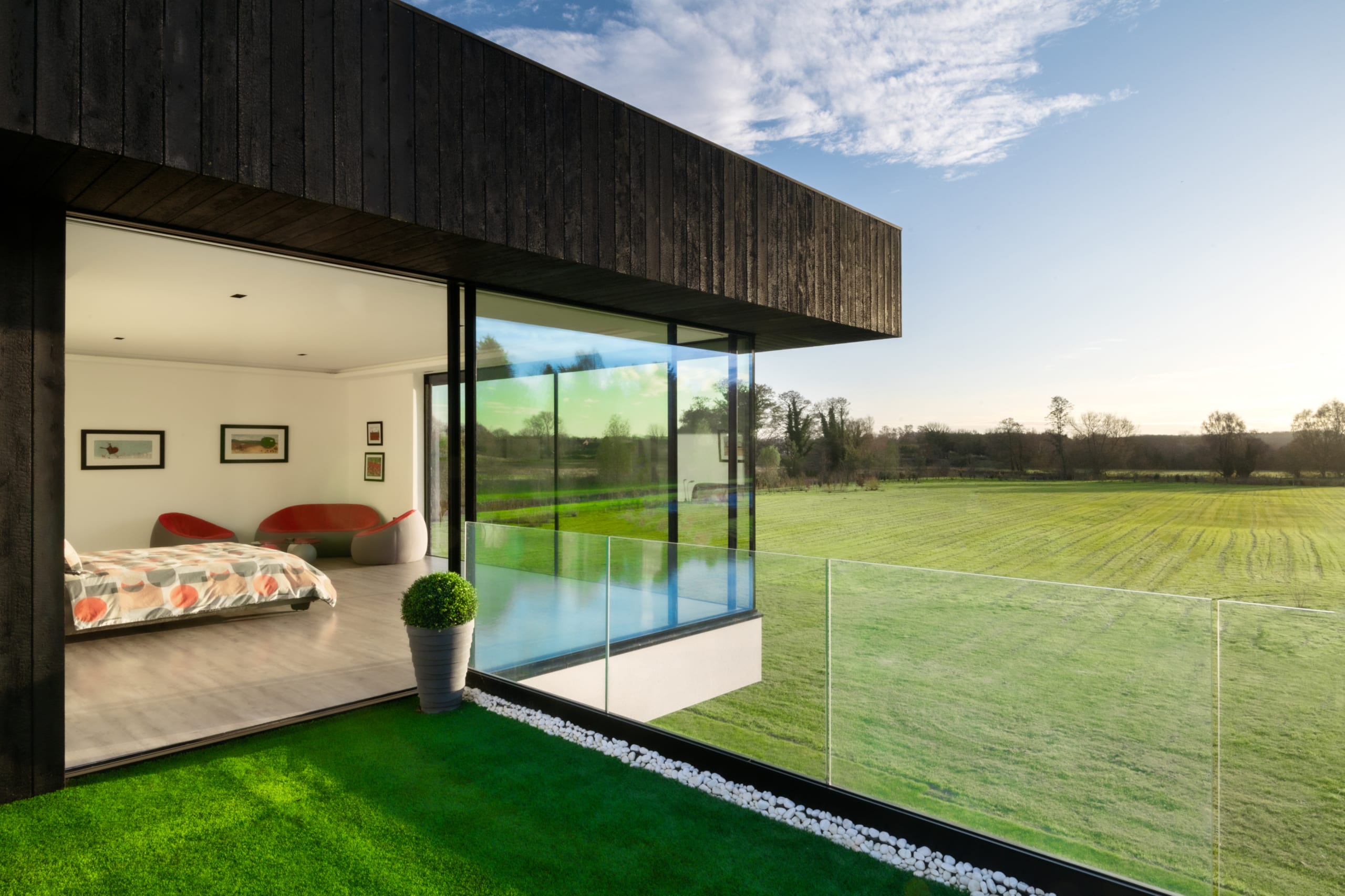 Modern bedroom with floor-to-ceiling glass windows opening to balcony overlooking green countryside, dark timber cladding exterior