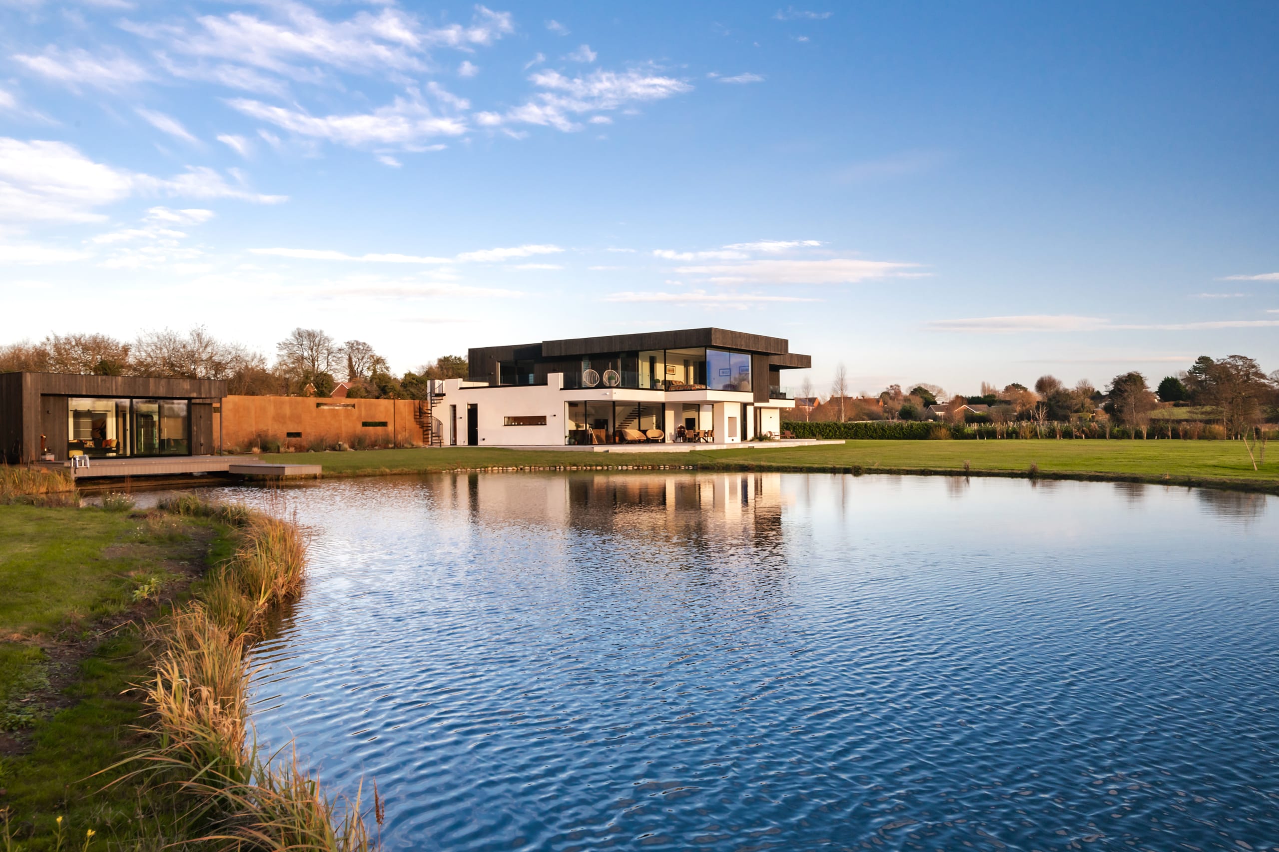 Modern two-story residence with dark upper volume and white lower level, featuring adjacent wood-clad pavilion beside a pond
