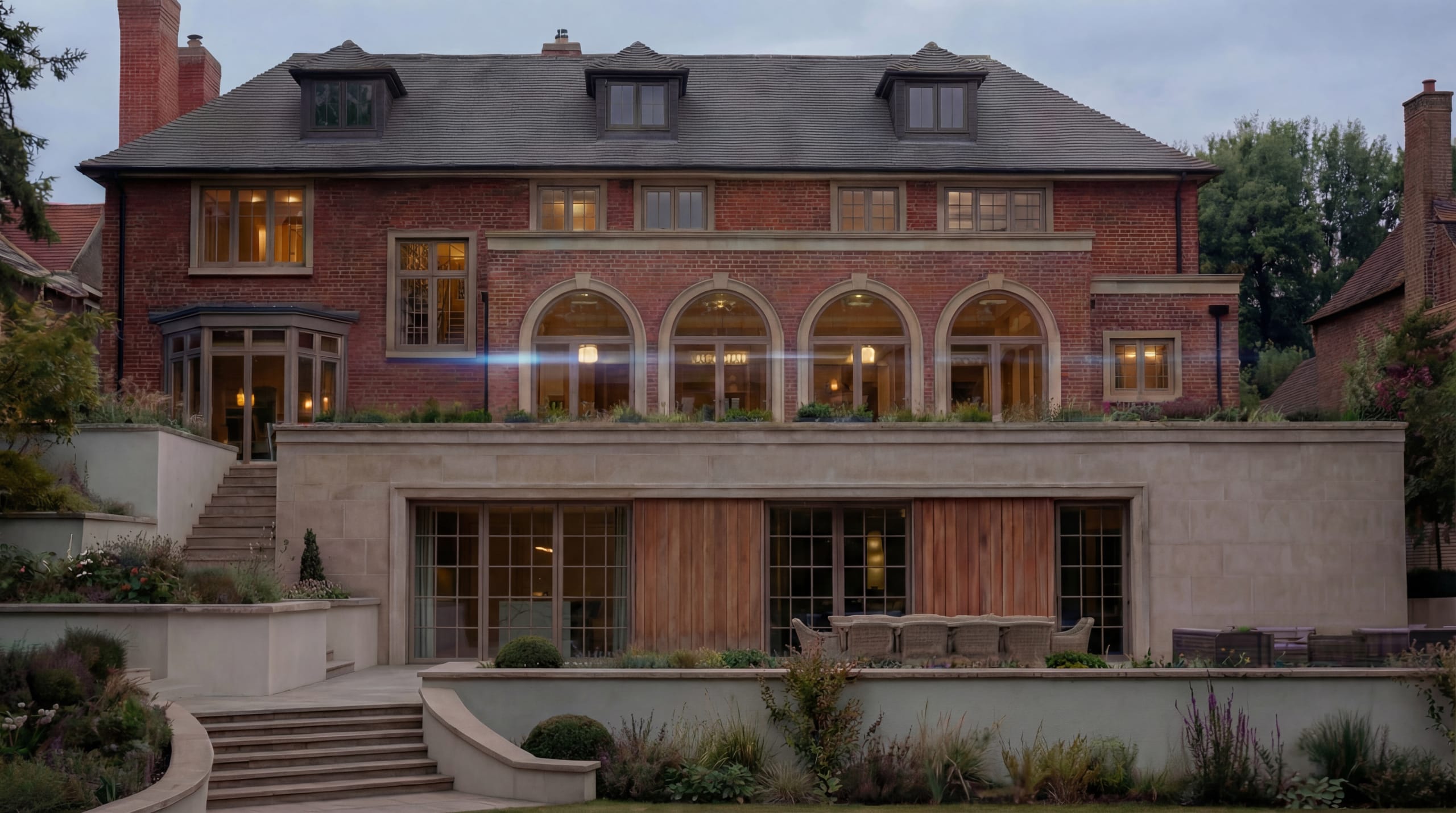 Elegant brick manor house with arched windows, stone terrace, and modern lower level extension at dusk