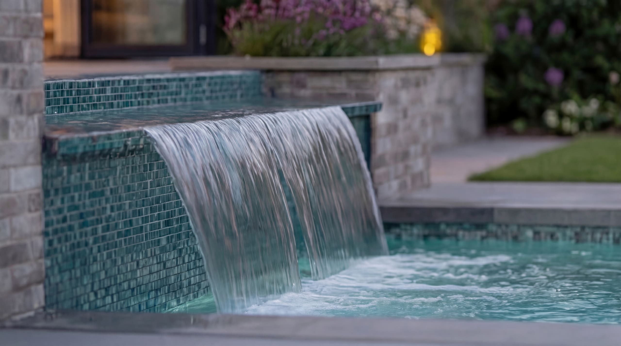 Water cascades over a green glass-tiled ledge into a backyard pool at dusk