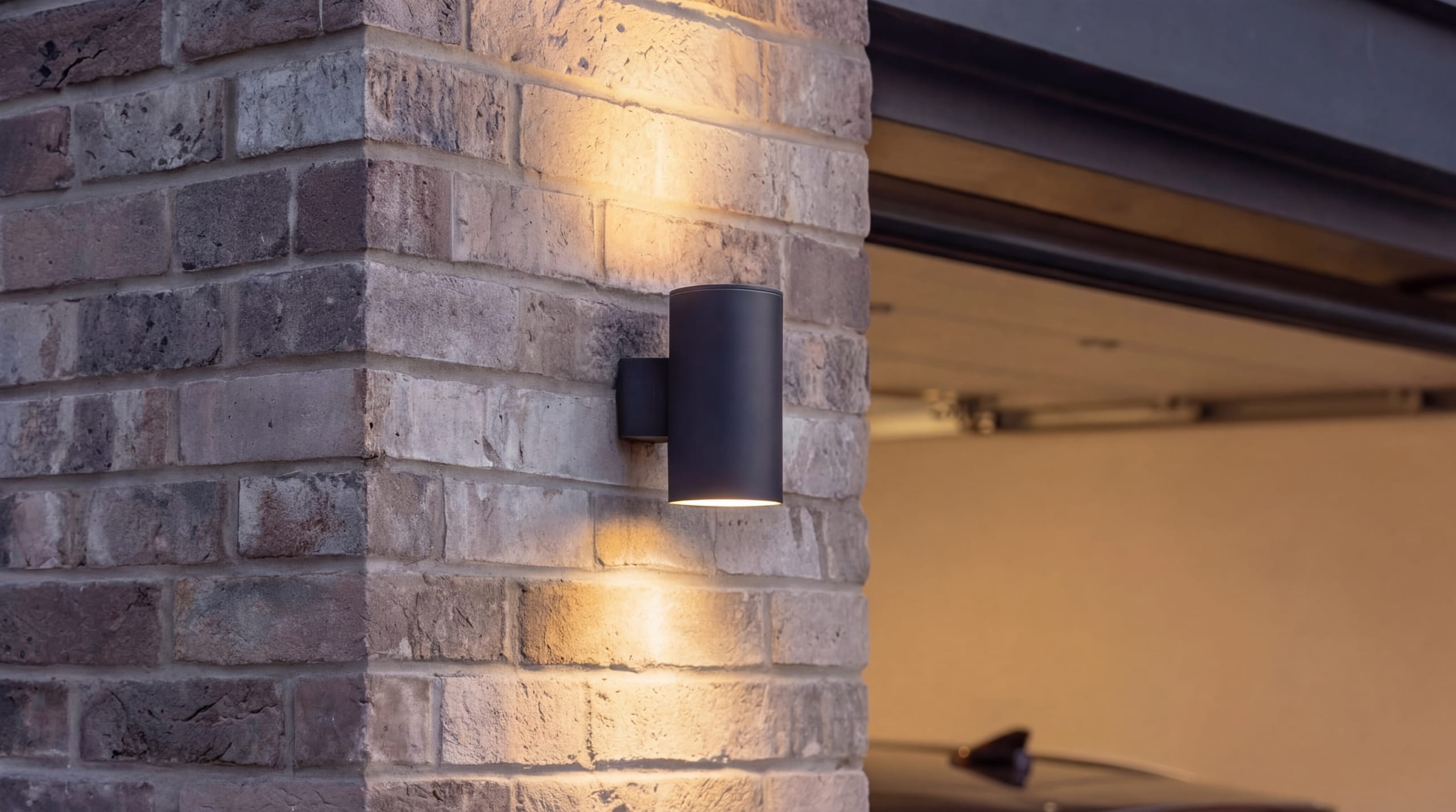 Cylindrical black up-down wall sconce mounted on textured brick corner beside an open garage