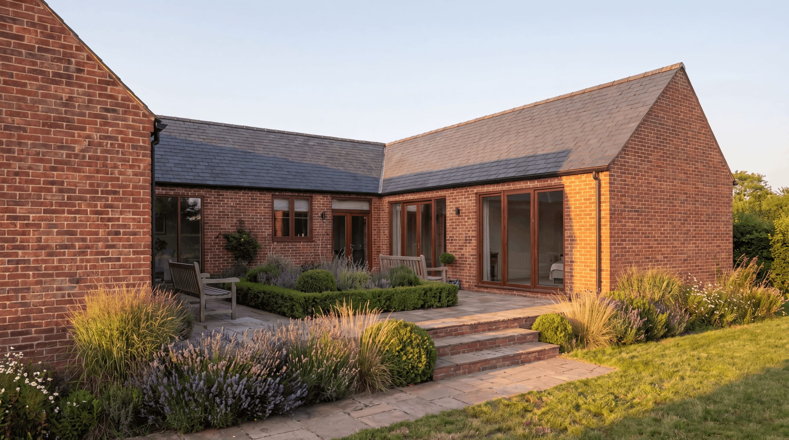 L-shaped brick single-storey house with slate roof, timber-framed doors, and a stone terrace bordered by grasses and lavender