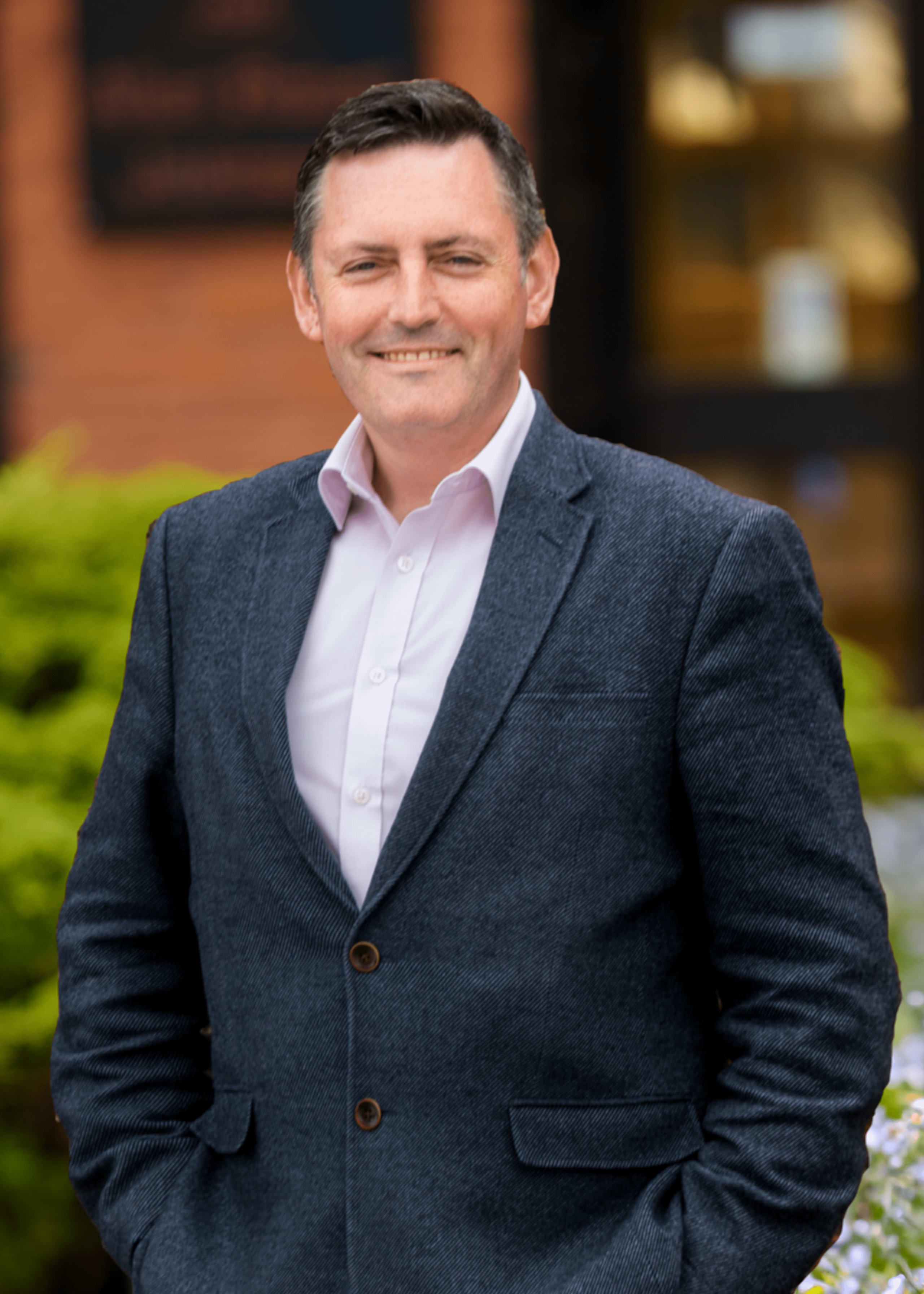 Smiling middle-aged man in charcoal blazer and white open-collar shirt standing outdoors in professional portrait