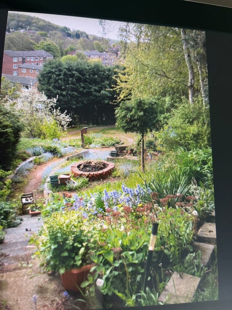 Lush terraced garden with circular brick fire pit, flowering plants, mature trees, and residential buildings in background