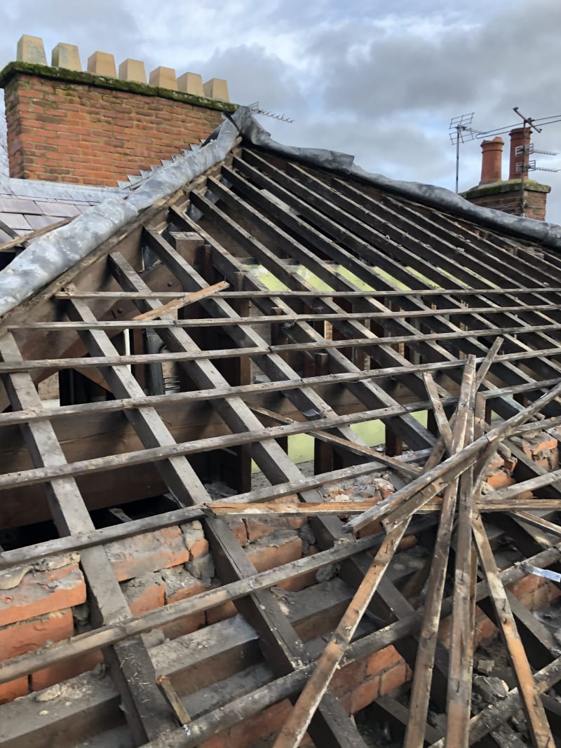 Stripped roof structure exposing timber rafters and battens with brick chimney stacks against a cloudy sky