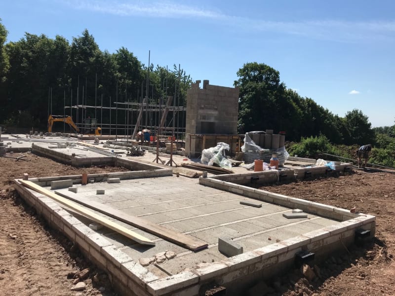 Construction site showing concrete slab foundations with block walls, scaffolding, and excavator amid rural tree-lined setting