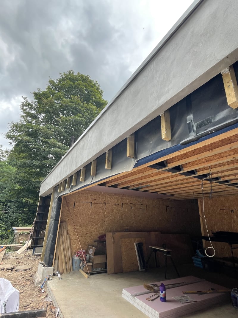 Modern building under construction showing exposed timber roof joists, OSB sheathing, and zinc or metal roof fascia detail