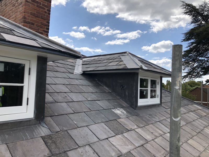 Slate-tiled roof with a white-framed dormer window and brick chimney under a partly cloudy sky