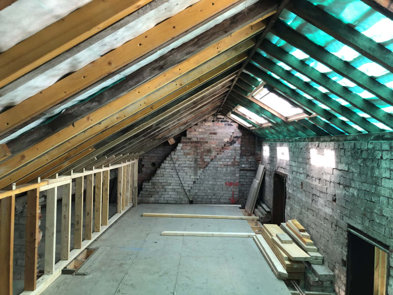 Loft conversion in progress showing exposed roof rafters, breathable membrane, and a skylight above a stripped brick gable wall