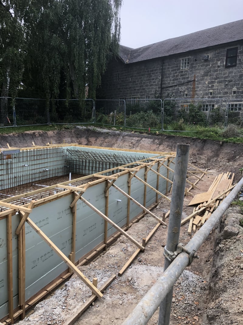 Excavated pool foundation with steel rebar grid and timber-braced formwork beside a stone barn