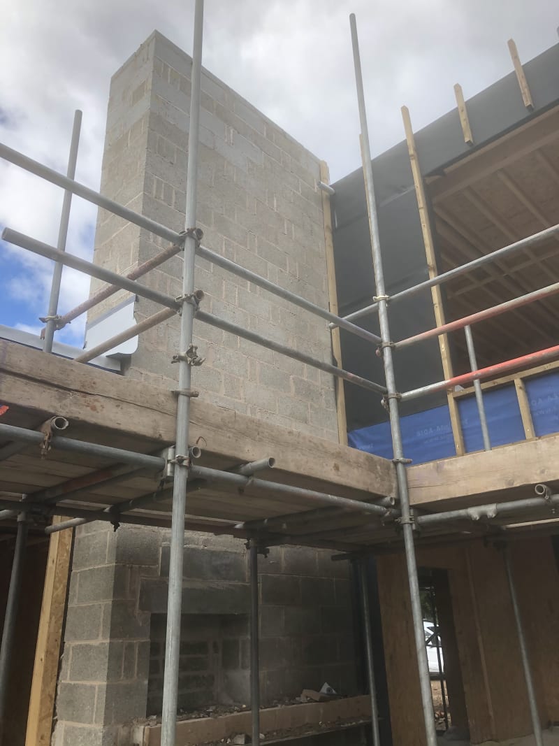 Concrete block chimney rising through a timber-framed house under construction, wrapped in scaffolding