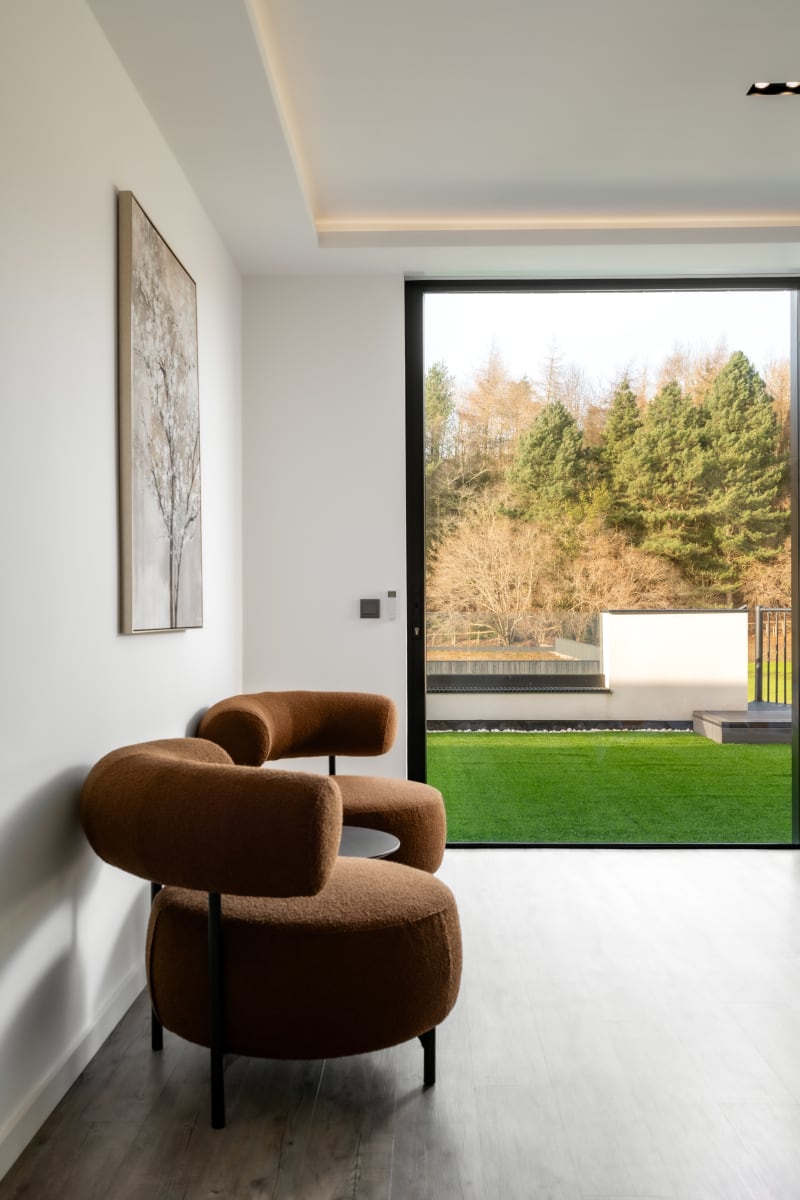 Modern interior with brown boucle armchair, floor-to-ceiling window overlooking manicured lawn and woodland landscape