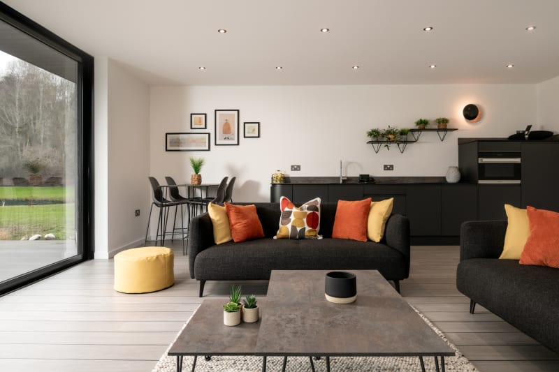 Modern open-plan living space with dark grey sofas, orange and yellow cushions, kitchenette, and large sliding glass doors