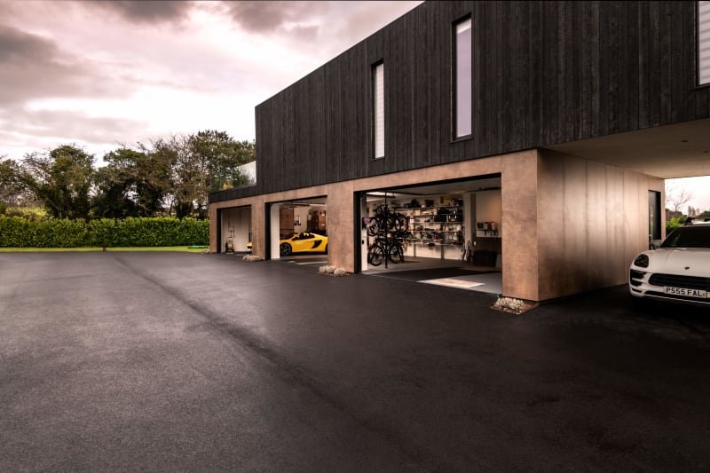 Modern two-story home with dark timber cladding, multi-bay garage featuring yellow McLaren and white Porsche sports cars