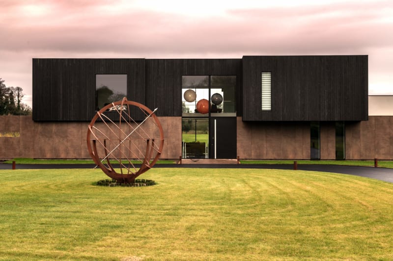 Modern two-story home with black timber and copper cladding, large windows, and spherical metal sculpture on manicured lawn
