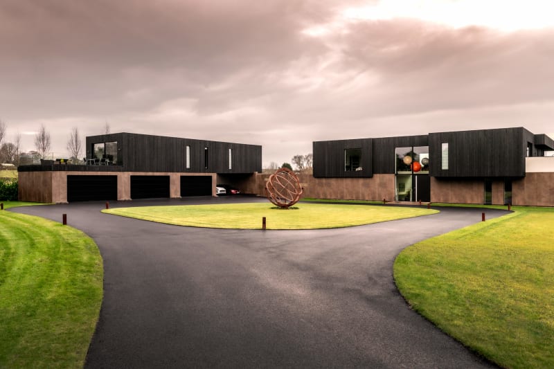 Modern black-clad residential complex with dual structures, circular driveway, manicured lawn, and spherical metal sculpture