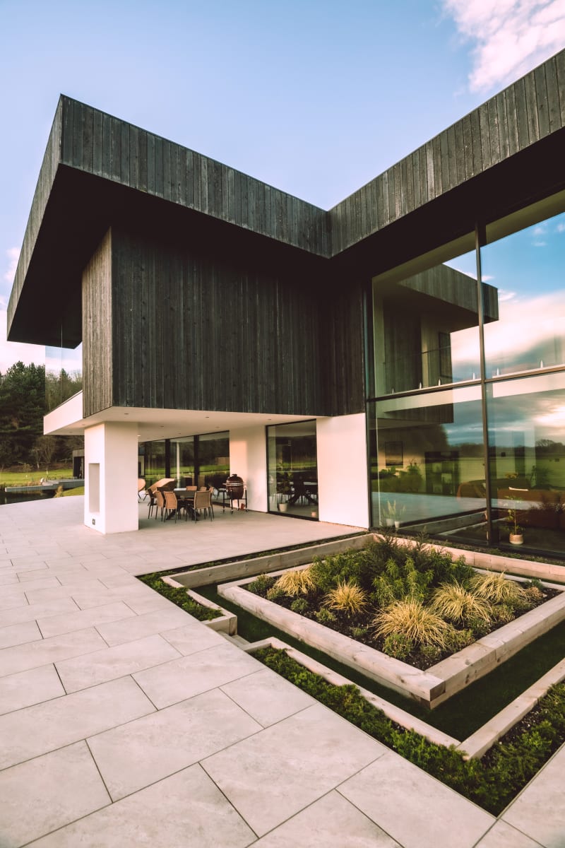 Modern two-story home with dark timber cladding, floor-to-ceiling windows, covered patio, and geometric raised garden beds