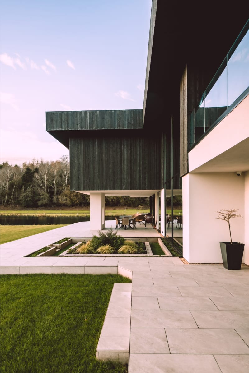 Modern lakeside home with dark timber cladding, white rendered walls, floor-to-ceiling glass, and covered outdoor terrace