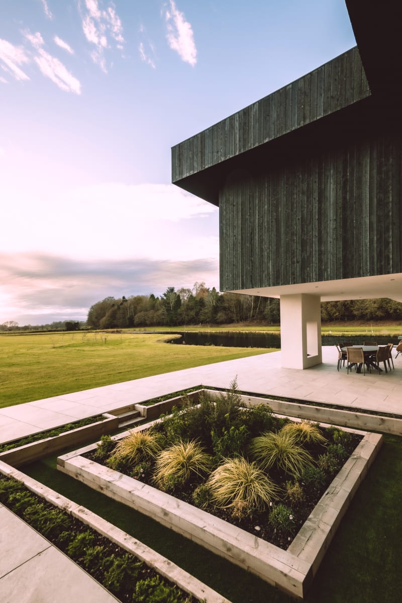 Modern cantilevered building with dark timber cladding over white base, sunken garden beds, patio dining area, and pastoral views
