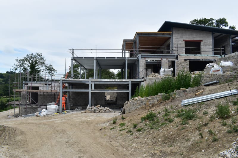 Modern residential construction site showing steel frame structure, concrete block walls, scaffolding, and stone retaining walls