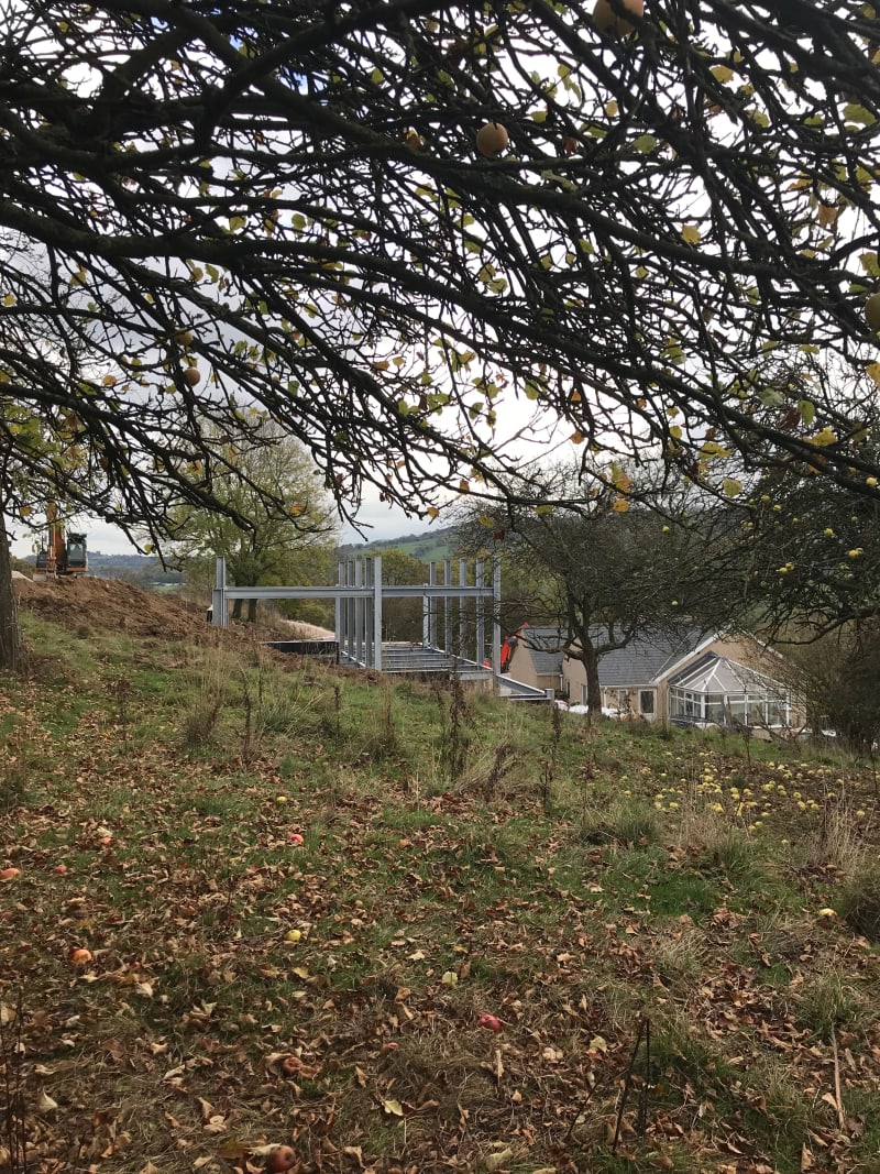 Steel frame structure under construction on hillside with existing house and conservatory, viewed through apple tree branches